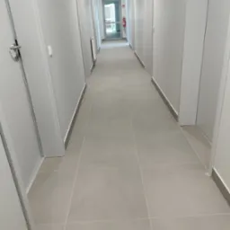 Długi, jasny korytarz z rzędem zamkniętych białych drzwi po obu stronach, wyłożony szarymi płytkami podłogowymi, z gaśnicą widoczną w oddali.