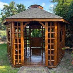 Mariusz Gruszczyński MG - Lakierowana, drewniana altana ogrodowa z ciemnoszarą, heksagonalną dachówką w słoneczny dzień, widok z zewnątrz na wypolerowaną podłogę i przylegający chodnik z kostki.