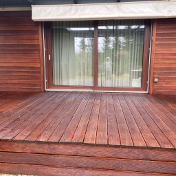 Mariusz Gruszczyński MG - Impregnowany taras z drewna egzotycznego, deski ułożone poziomo, widoczne słoje drewna, brązowa kolorystyka, w tle przesuwne drzwi tarasowe i fragment elewacji.
