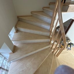 Mariusz Gruszczyński MG - Jasne, drewniane schody spiralne z balustradą, widok z góry. Widoczne narzędzia i materiały budowlane w tle. Naturalne oświetlenie podkreśla strukturę drewna.