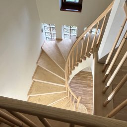 Mariusz Gruszczyński MG - Drewniane, kręte schody z jasnego drewna, widziane z góry. Stopnie wykończone, balustrada z okrągłych elementów. W tle okna z luksferami i małe okno.