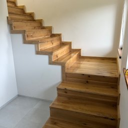 Mariusz Gruszczyński MG - Drewniane schody dębowe, lakierowane, z widocznymi słojami drewna, zamontowane przy białej ścianie. Stopnie schodów o nieregularnym kształcie, widok z dołu.