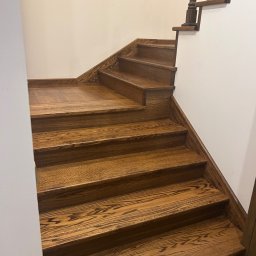 Mariusz Gruszczyński MG - Odnowione drewniane schody z ciemnym bejcowaniem, widoczne słoje drewna. Ujęcie z dołu, ukazujące detale wykończenia i kontrast z białą ścianą.