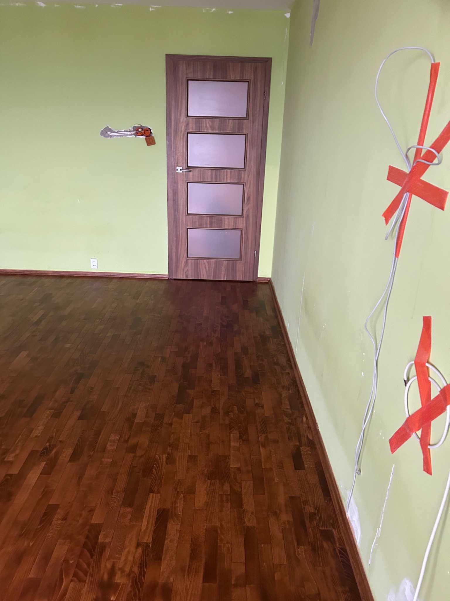 Pokój w trakcie remontu: świeżo położona, ciemna podłoga z parkietu, ściany w odcieniach zieleni z widocznymi śladami prac, zamontowane drzwi z drewnianą ramą i szybami, wystające przewody...