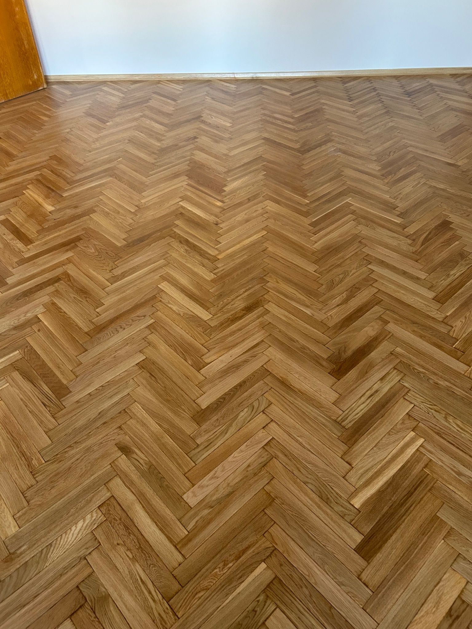 Ułożona w jodełkę, lakierowana, parkietowa podłoga z drewna dębowego, widok z góry, fragment drzwi w lewym górnym rogu.