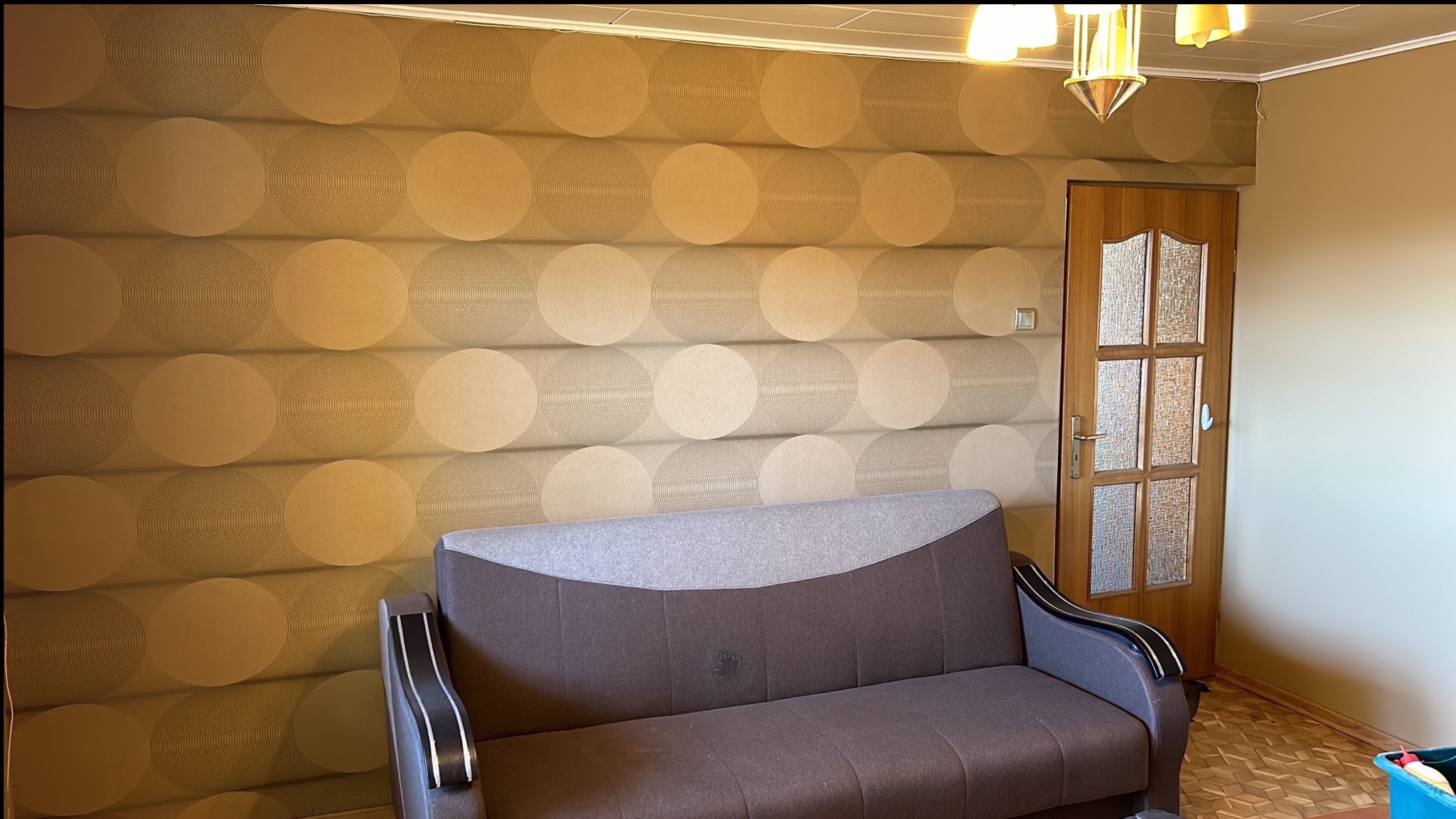 Wnętrze pokoju z brązową sofą na tle ściany pokrytej tapetą z geometrycznym wzorem w odcieniach beżu i brązu, obok drewniane drzwi z przeszkleniami.