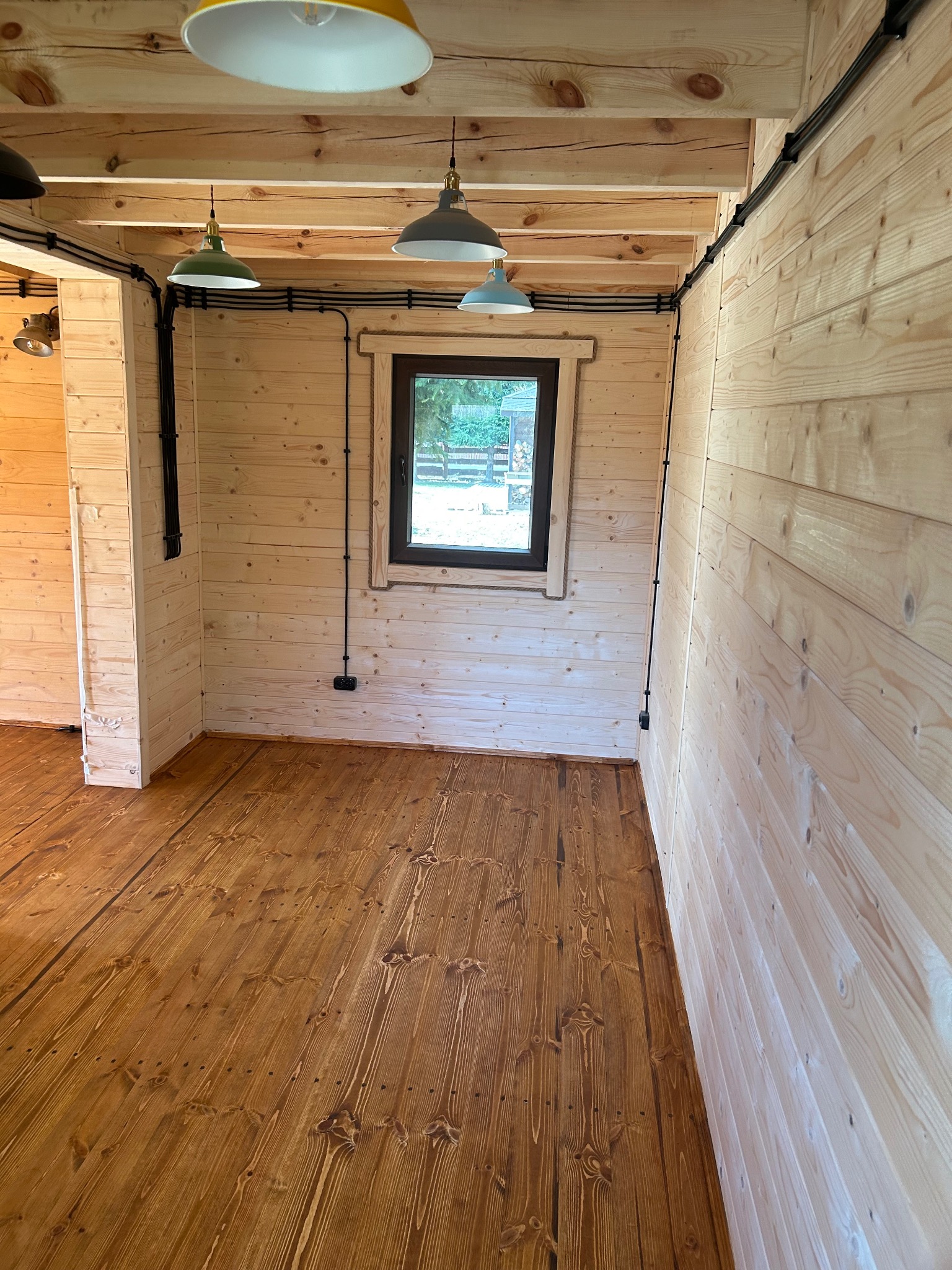 Wnętrze drewnianego domu z oknem, widoczne pomalowane drewniane ściany, podłoga po cyklinowaniu i lakierowaniu, oraz instalacja elektryczna natynkowa z lampami w stylu retro.