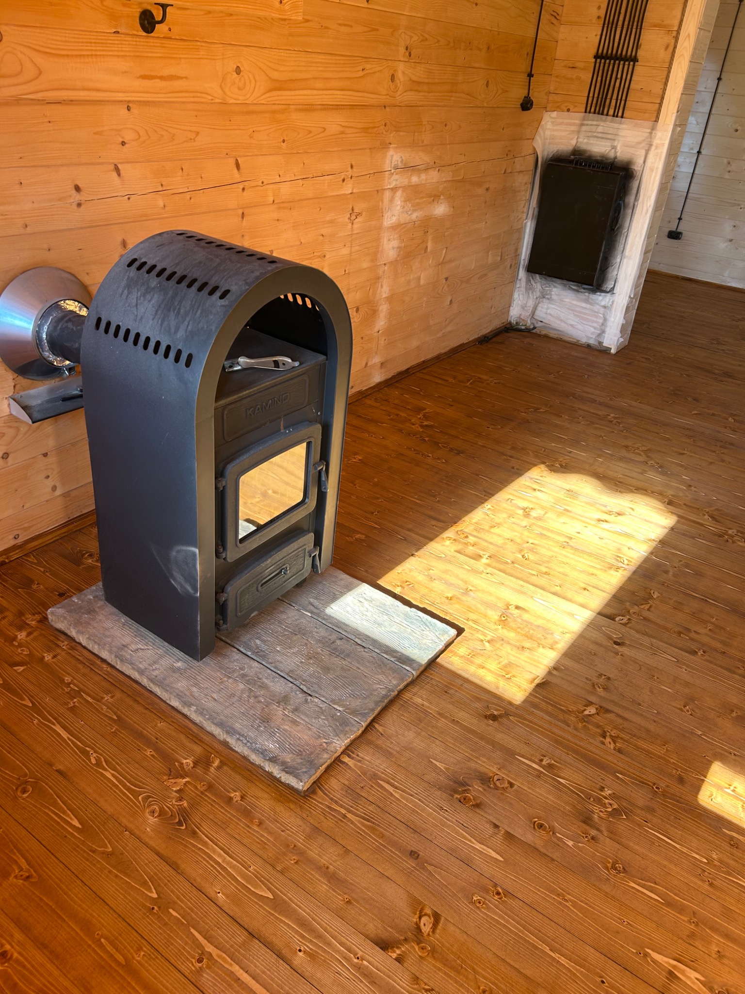 Wnętrze drewnianego domu z zainstalowanym czarnym piecem kominkowym na drewnianej podłodze, widoczne promienie słoneczne padające na podłogę i obudowa komina w tle.