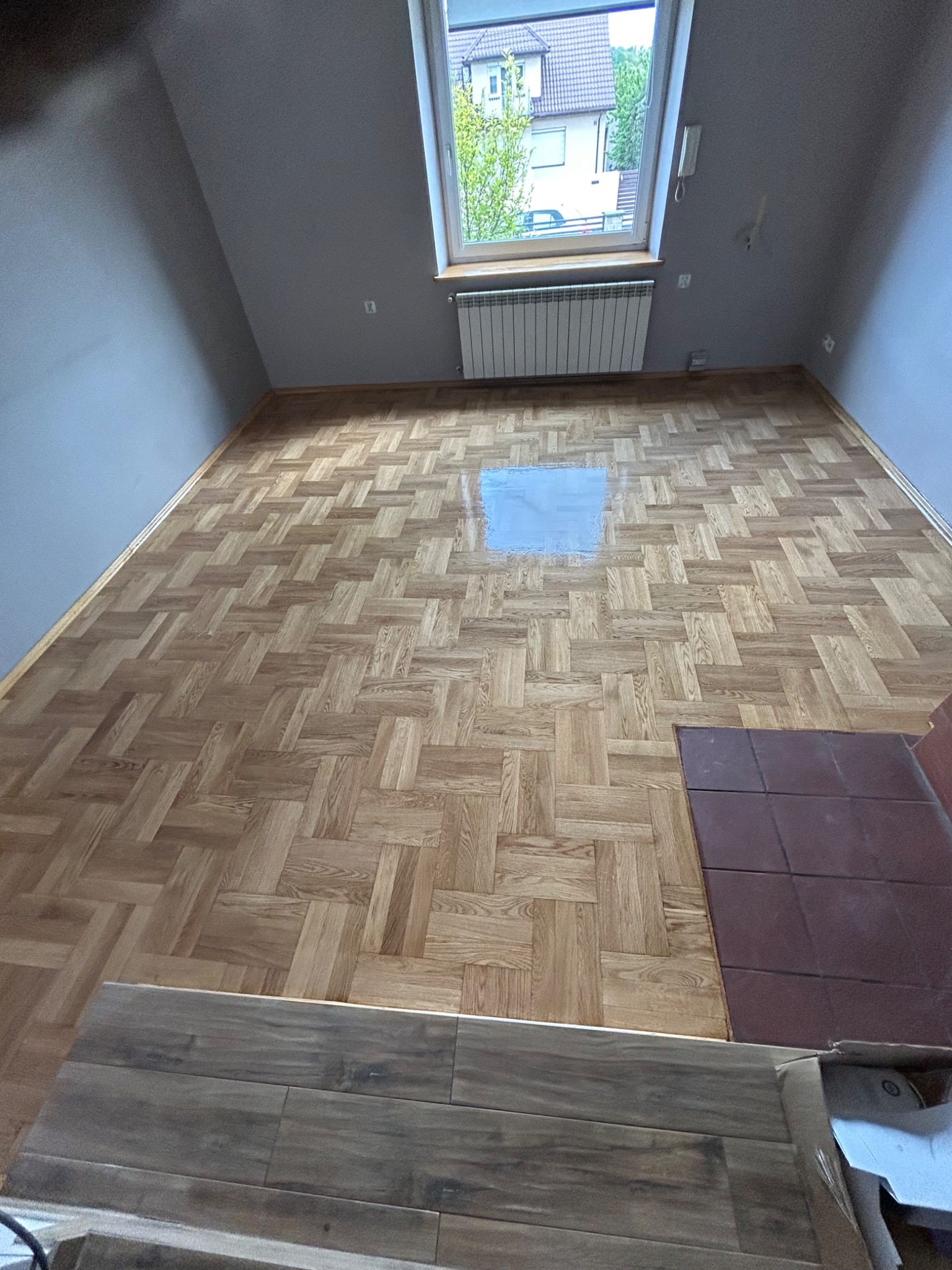 Parkiet w jodełkę po cyklinowaniu, z widocznym fragmentem podkładu, okno dachowe i szare ściany. Część podłogi zakryta nowymi panelami.