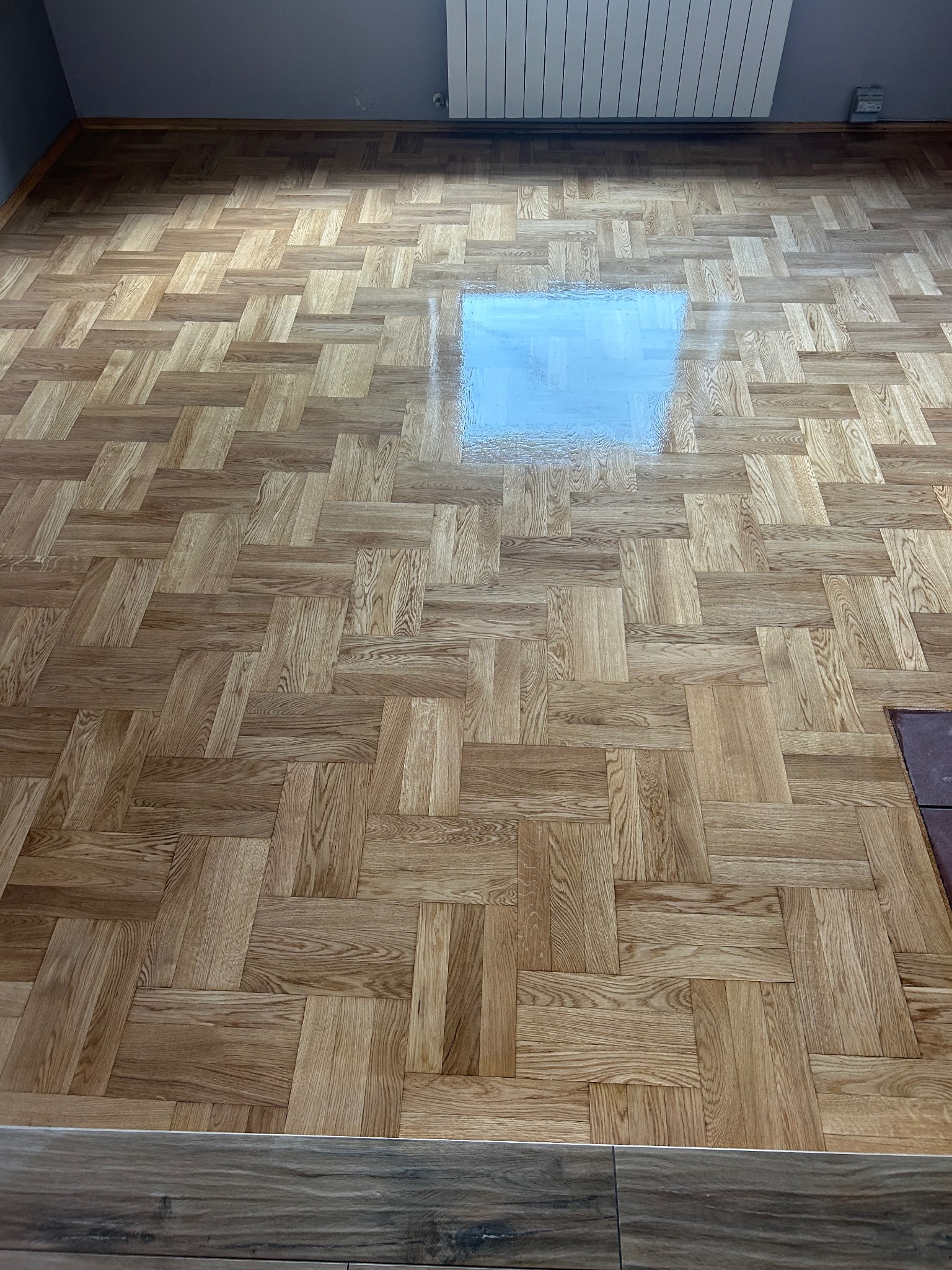 Parkiet ułożony w jodełkę, po cyklinowaniu, z widocznym połyskiem od lakieru. Fragment ściany i grzejnika w tle, podkreślający wnętrze. Jasne drewno.