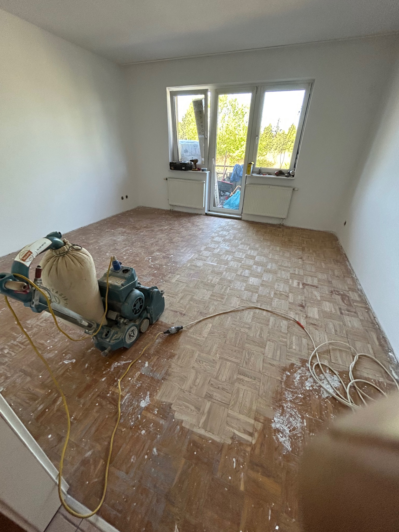 Cyklinowanie parkietu maszyną w pokoju z oknem balkonowym. Widoczne ślady pyłu po szlifowaniu, ukazujące postęp prac renowacyjnych podłogi drewnianej.