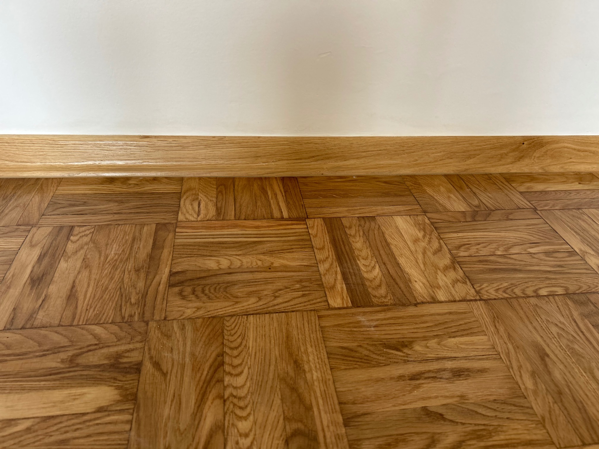 Parkiet w jodełkę z listwą przypodłogową z jasnego drewna na tle białej ściany. Widok z góry, ukazujący detale drewnianej podłogi i jej wzór.