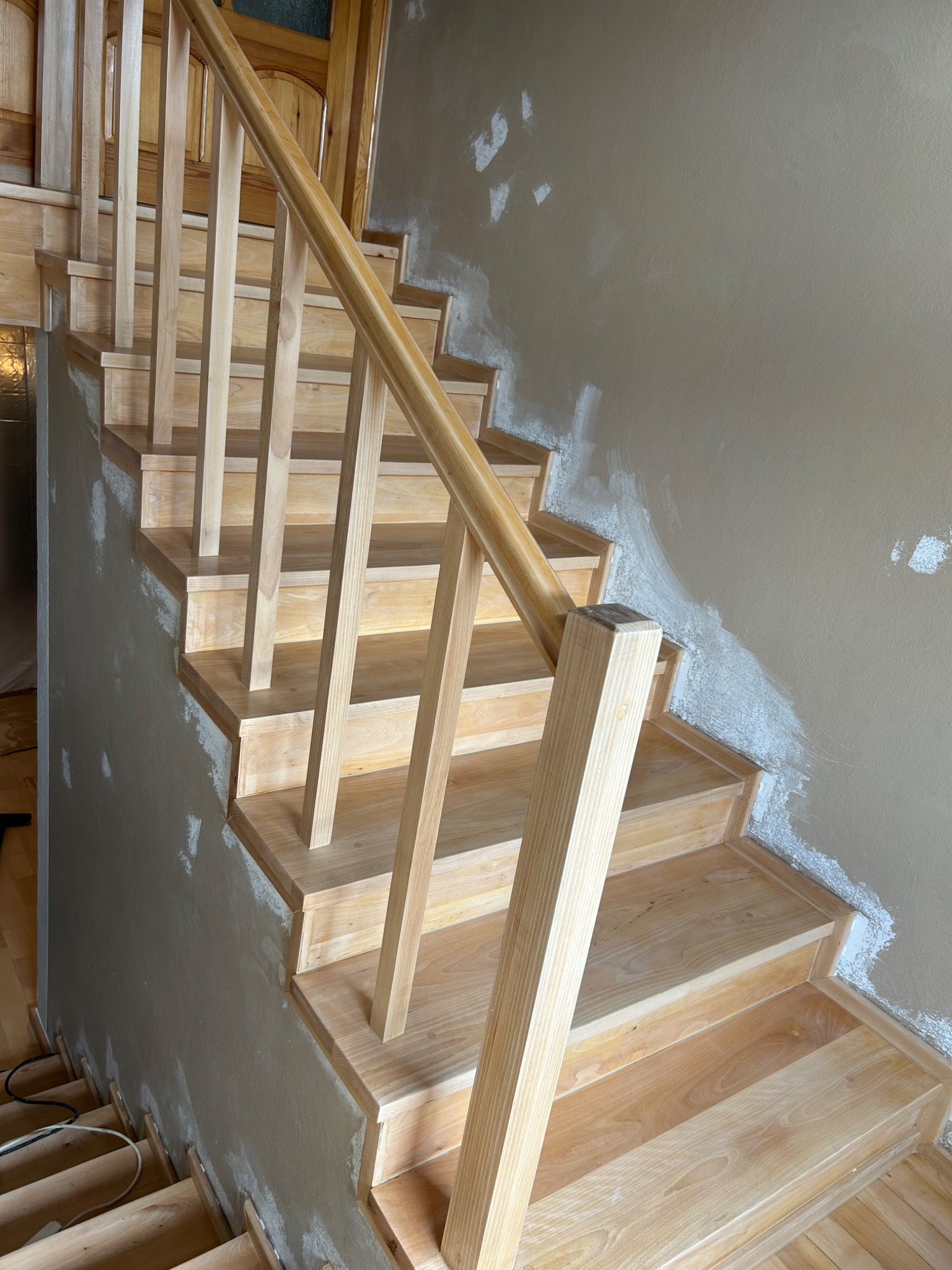 Jasne, drewniane schody z balustradą w trakcie montażu. Ściana przygotowana pod malowanie z widocznymi śladami szpachlowania. Widok z dołu na górę.