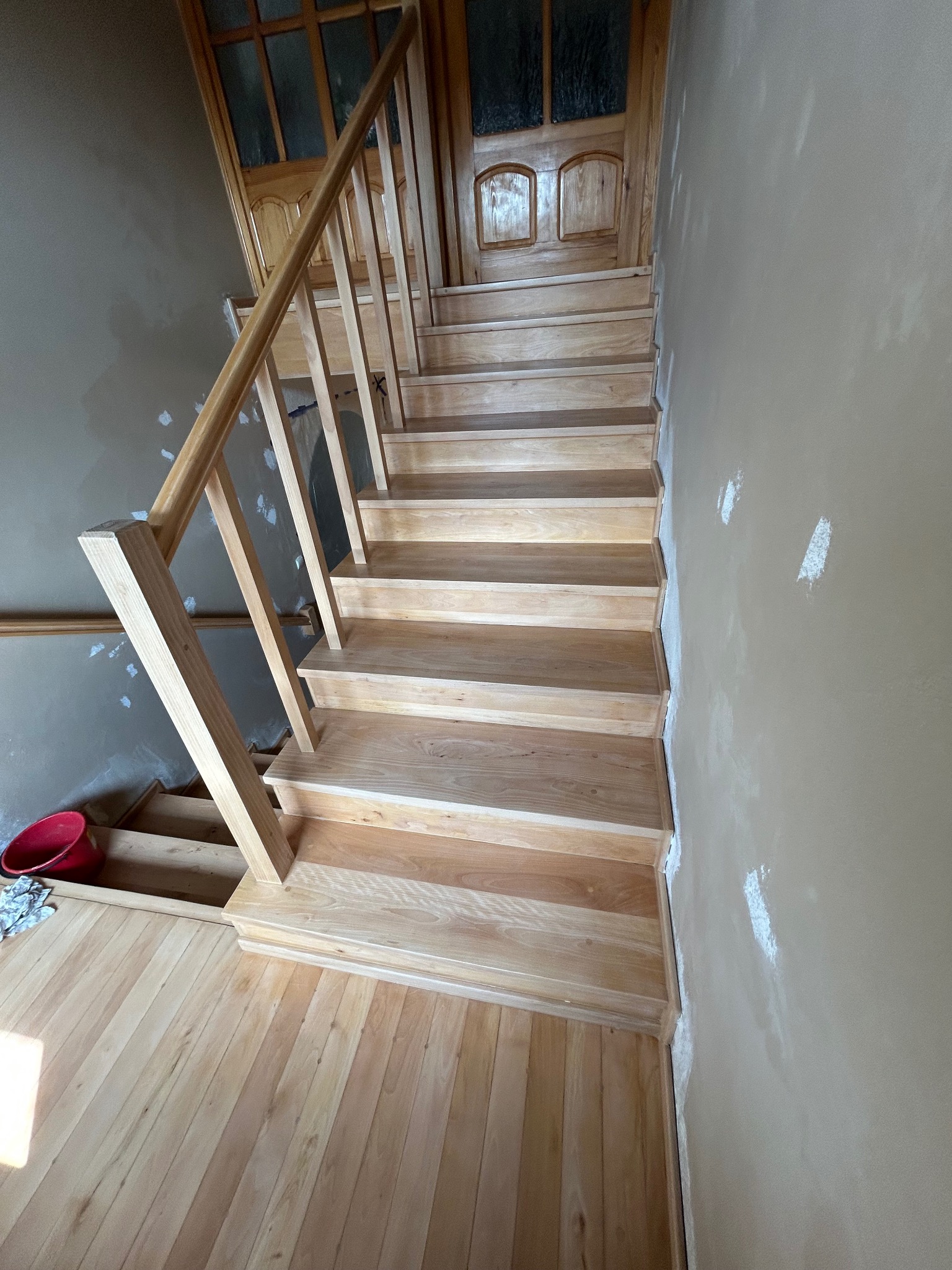 Drewniane schody z jasnego drewna, prowadzące do góry, widoczne w Rudnikach. Ściany przygotowane do malowania, drewniane wykończenie podłogi i drzwi.