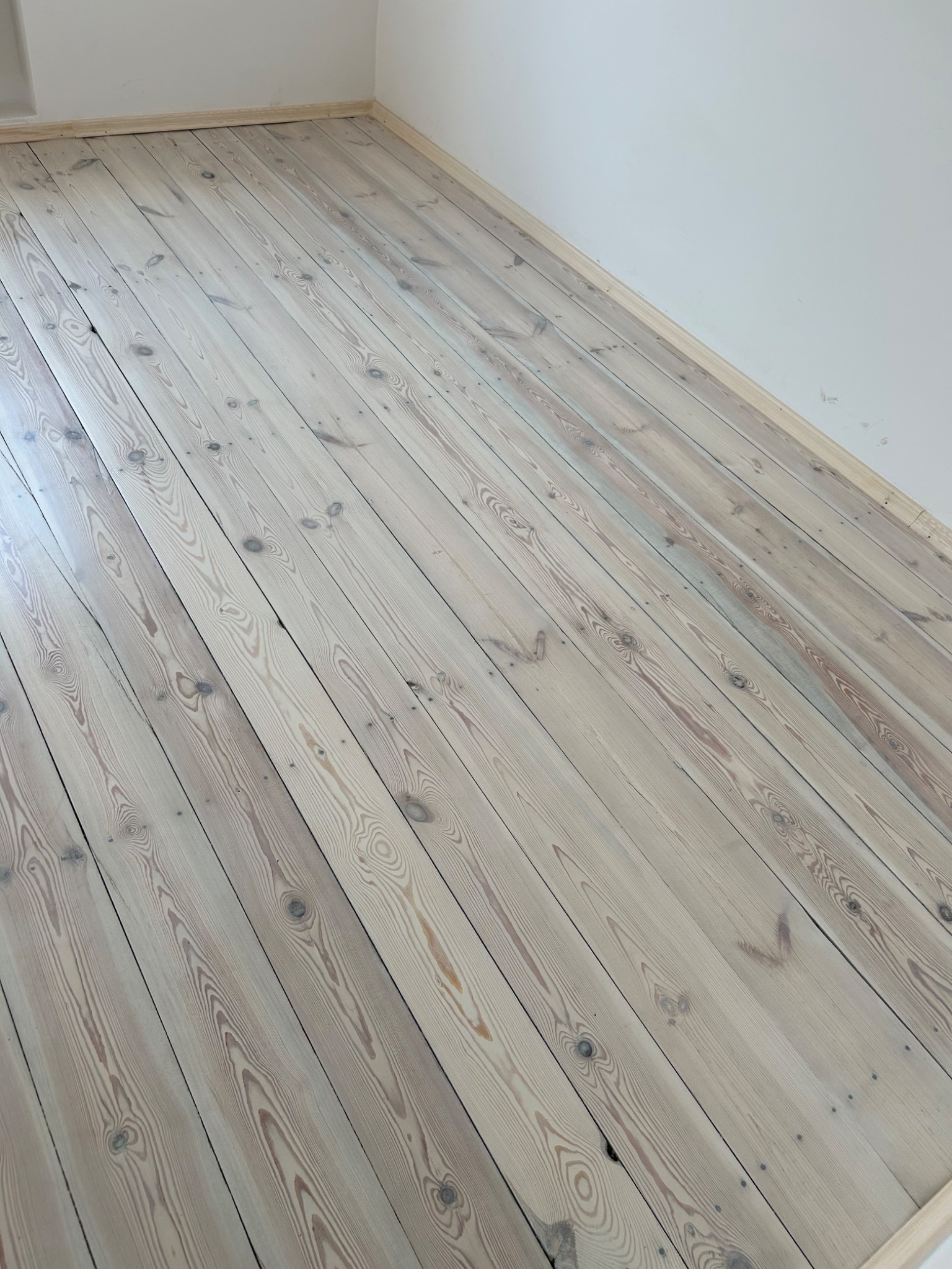 Jasne, odnowione deski podłogowe z widocznym usłojeniem, ułożone ukośnie w pustym, pomalowanym na biało pokoju z listwami przypodłogowymi.