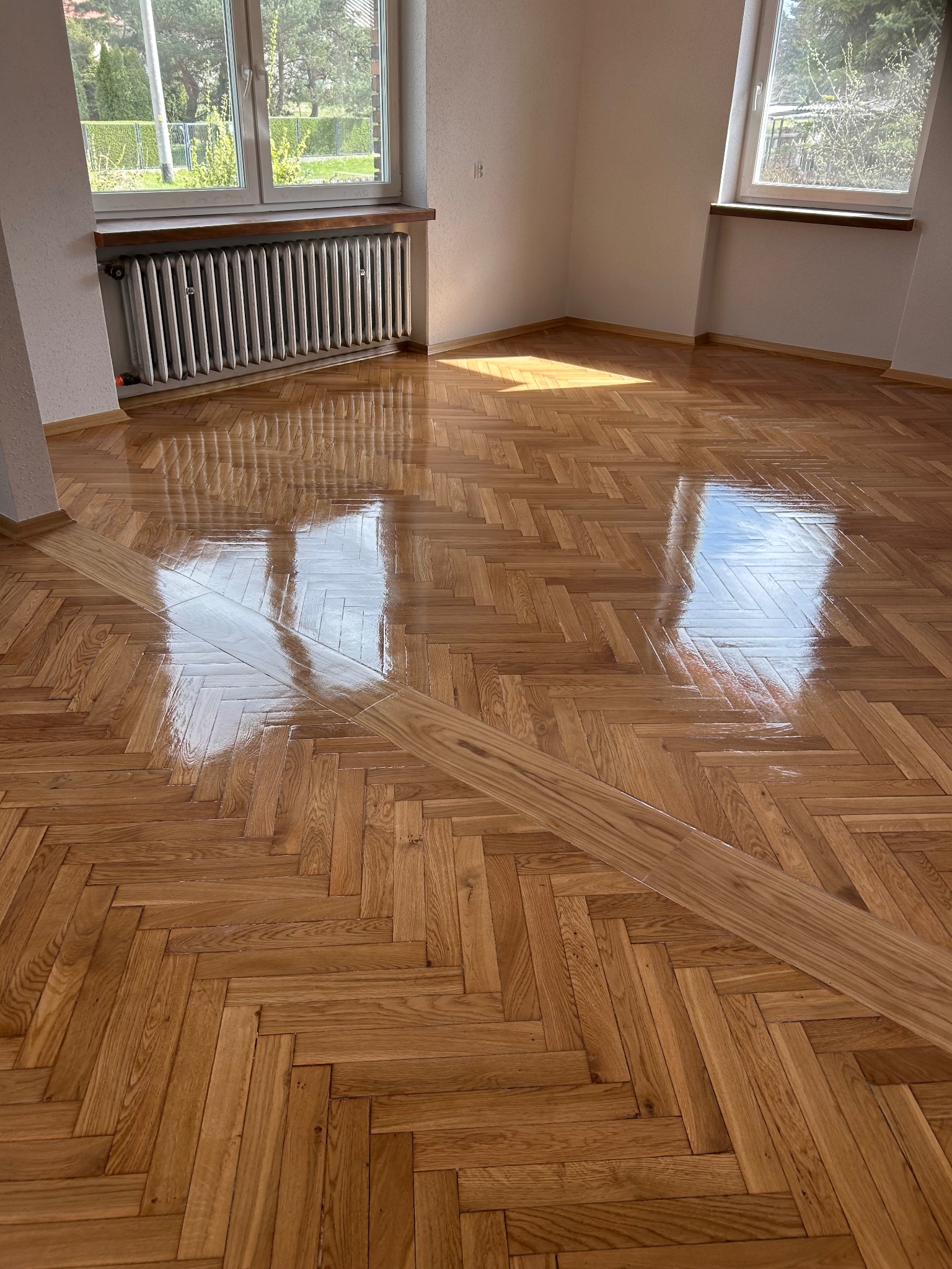 Błyszcząca, odnowiona parkietowa podłoga w jodełkę po cyklinowaniu, z wyraźnym wzorem drewna i refleksami światła w pustym pokoju z oknami.