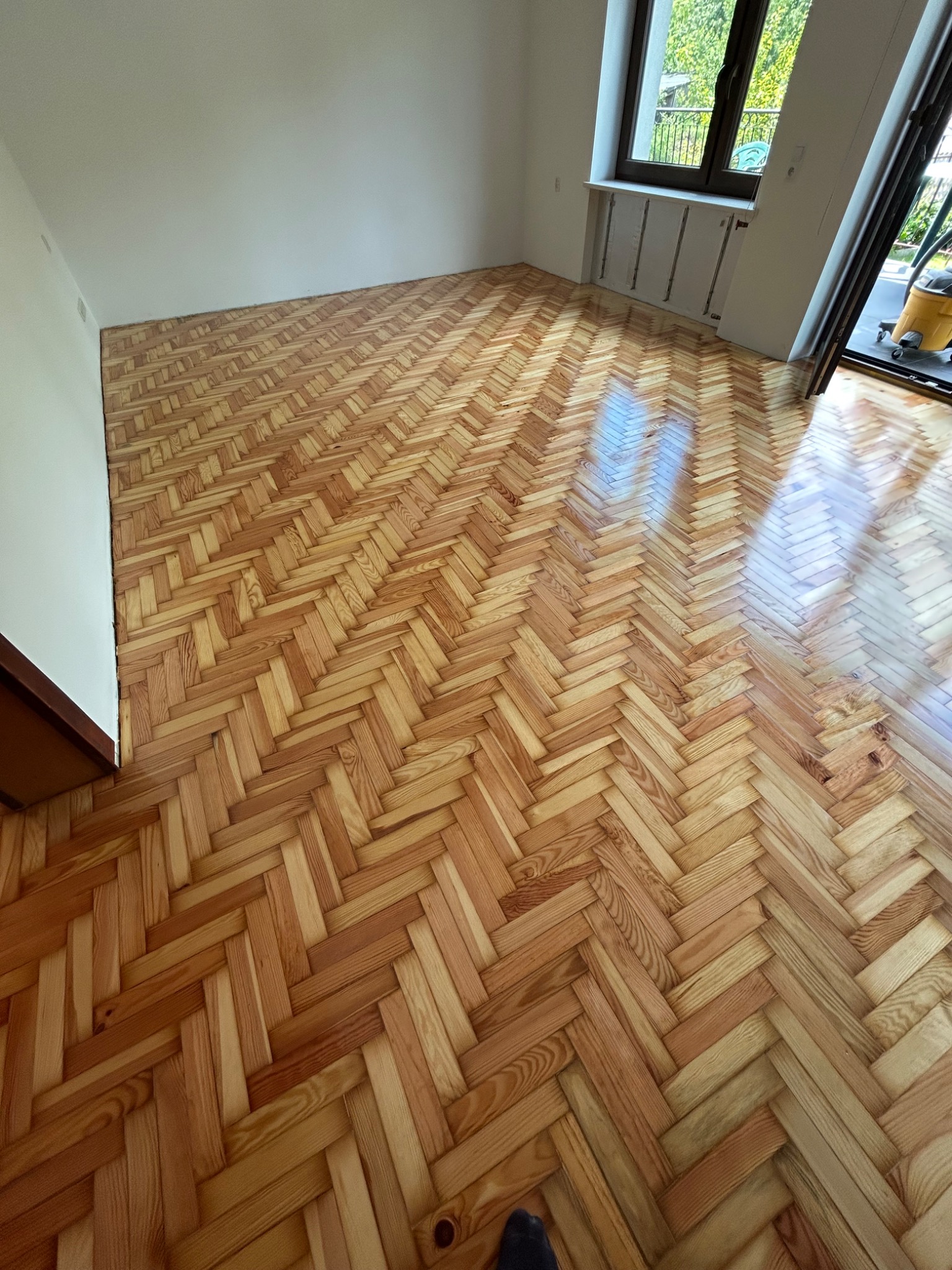 Parkiet ułożony w jodełkę, po cyklinowaniu, z widocznym oknem i elementami wyposażenia. Jasne drewno, świeżo po renowacji, odbija światło.