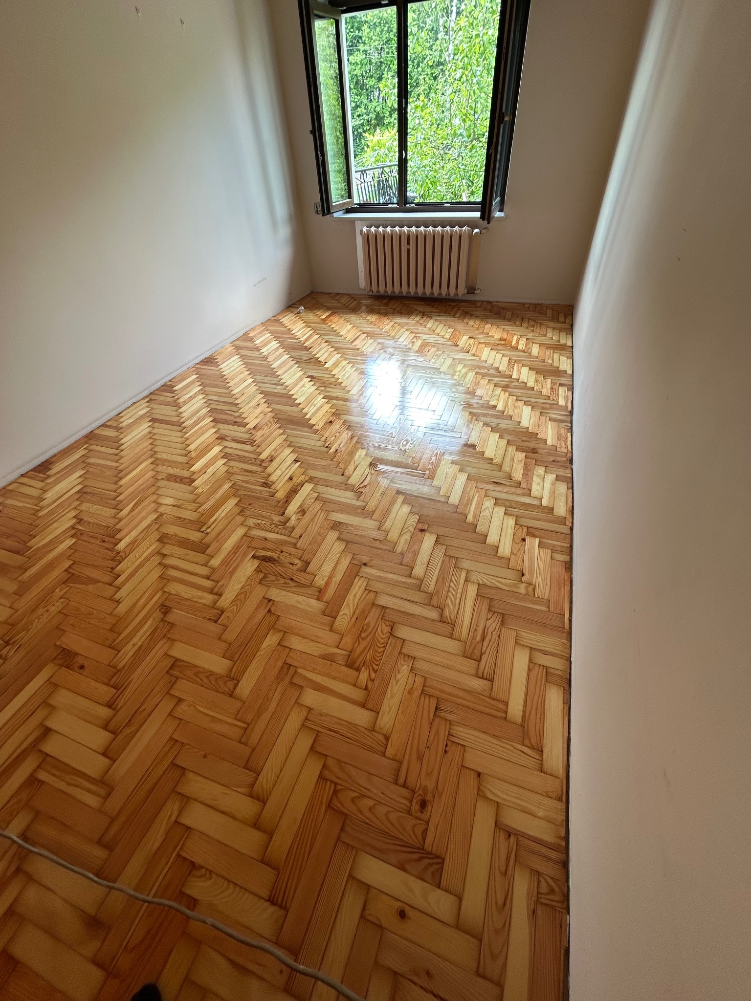 Parkiet ułożony w jodełkę w pustym pokoju z oknem i grzejnikiem. Świeżo położona, błyszcząca podłoga odbija światło. Widok na zieleń za oknem.