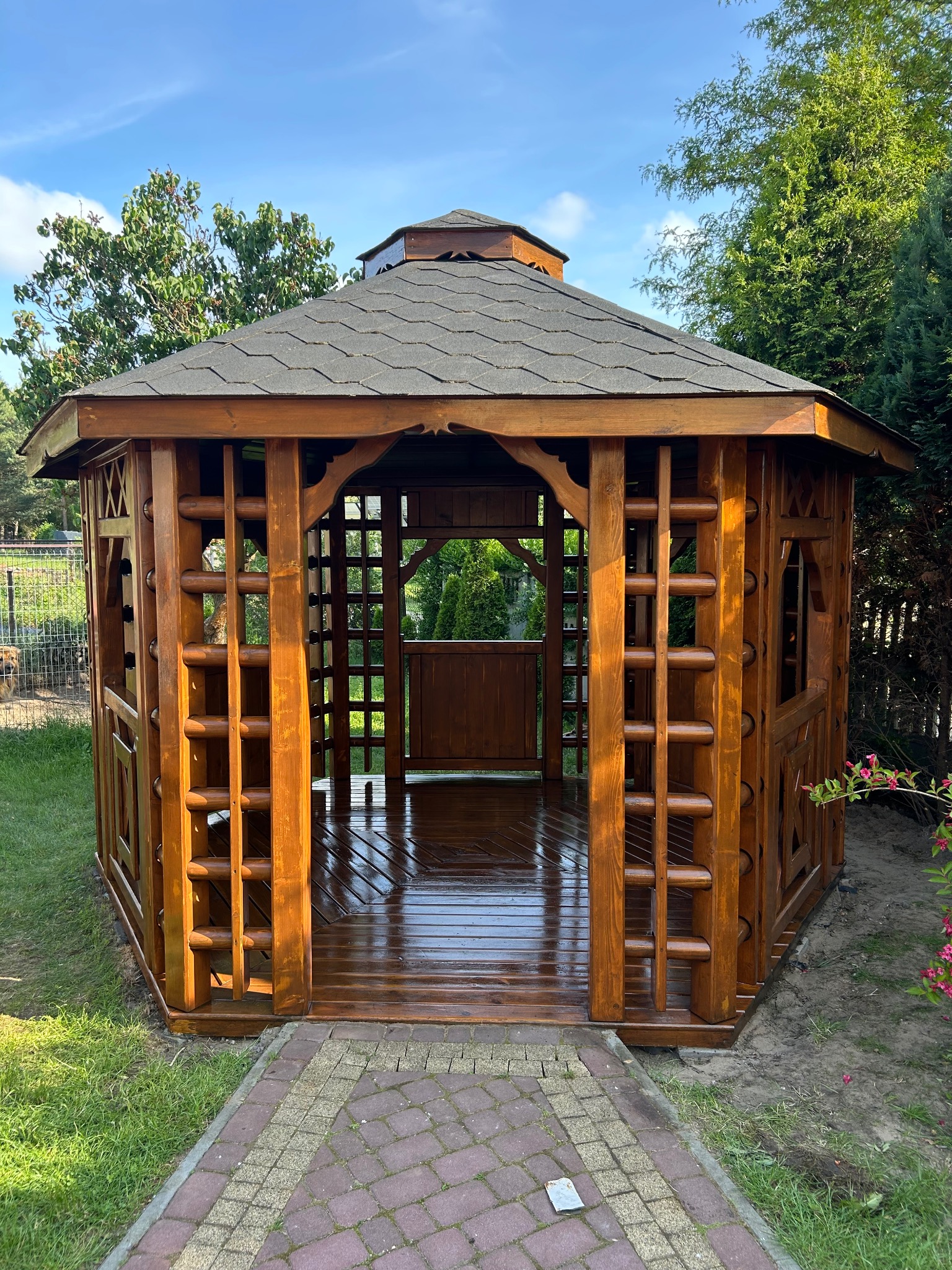 Lakierowana, drewniana altana ogrodowa z ciemnoszarą, heksagonalną dachówką w słoneczny dzień, widok z zewnątrz na wypolerowaną podłogę i przylegający chodnik z kostki.