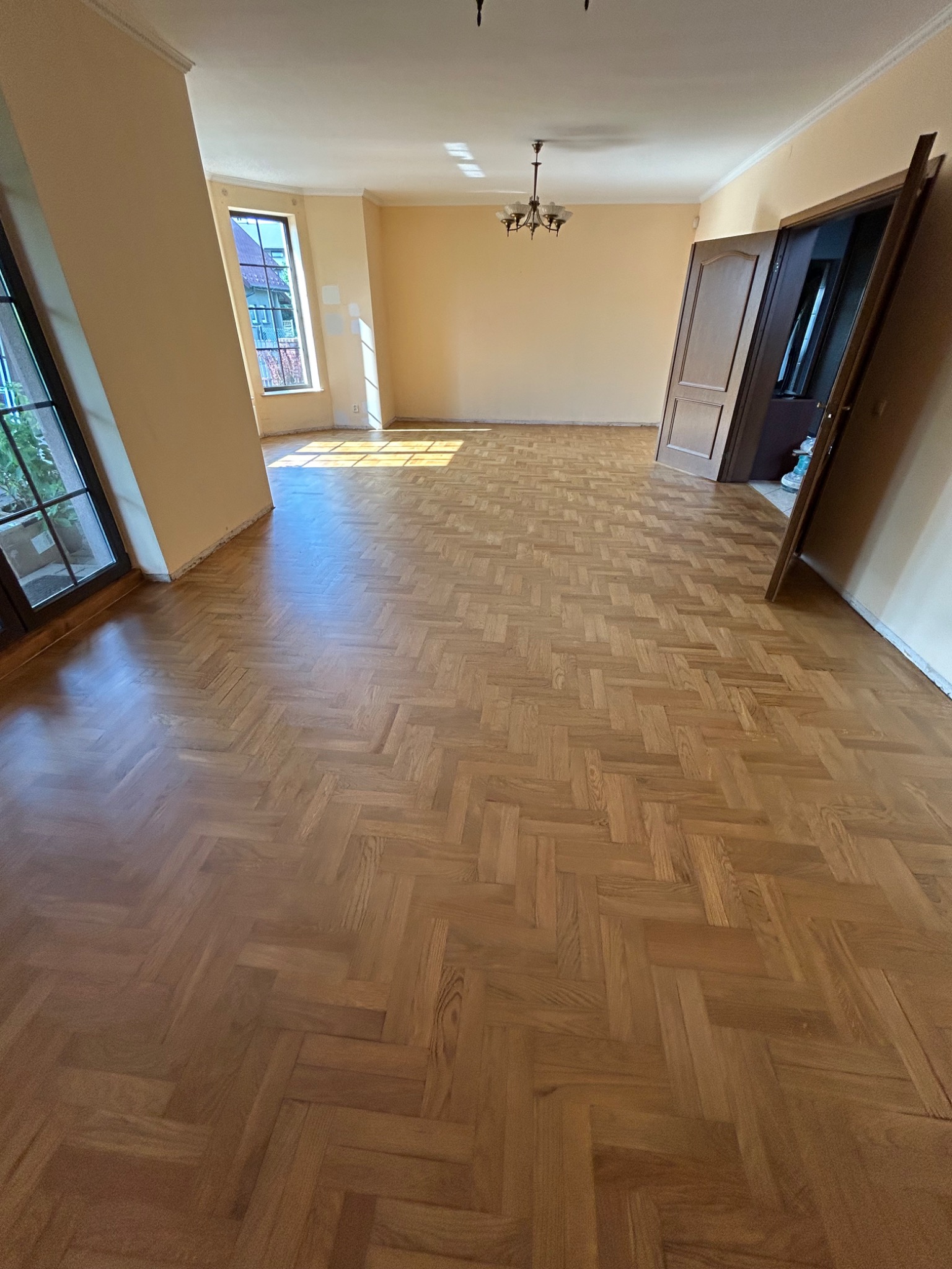 Salon z parkietem ułożonym w jodełkę po cyklinowaniu. Widoczne drzwi, okno z widokiem na zewnątrz. Świeże wykończenie i jasne, słoneczne wnętrze.