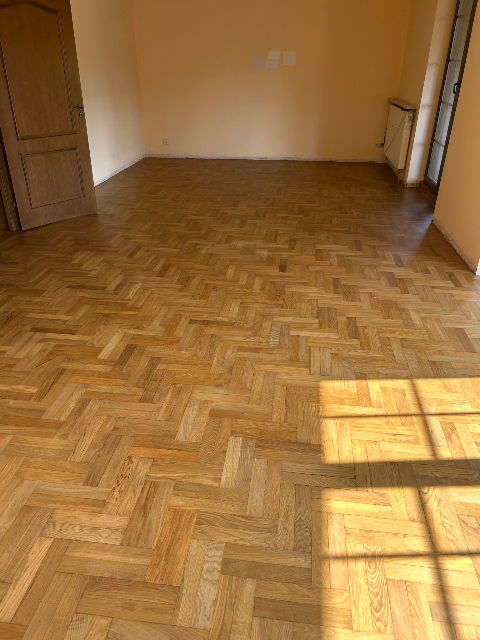 Parkiet ułożony w jodełkę w pustym pokoju z beżowymi ścianami. Widoczne drzwi, grzejnik i cień okna. Podłoga po cyklinowaniu, z wyraźnym rysunkiem drewna i połyskiem.