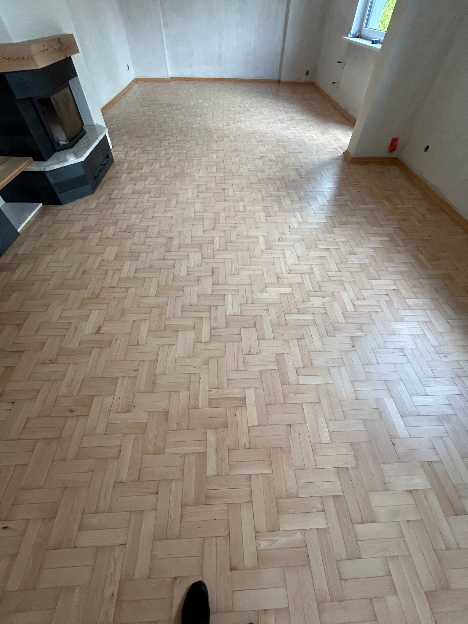 Jasna, parkietowa podłoga ułożona w jodełkę francuską, świeżo po cyklinowaniu, z widocznym kominkiem po lewej stronie i oknem w tle.