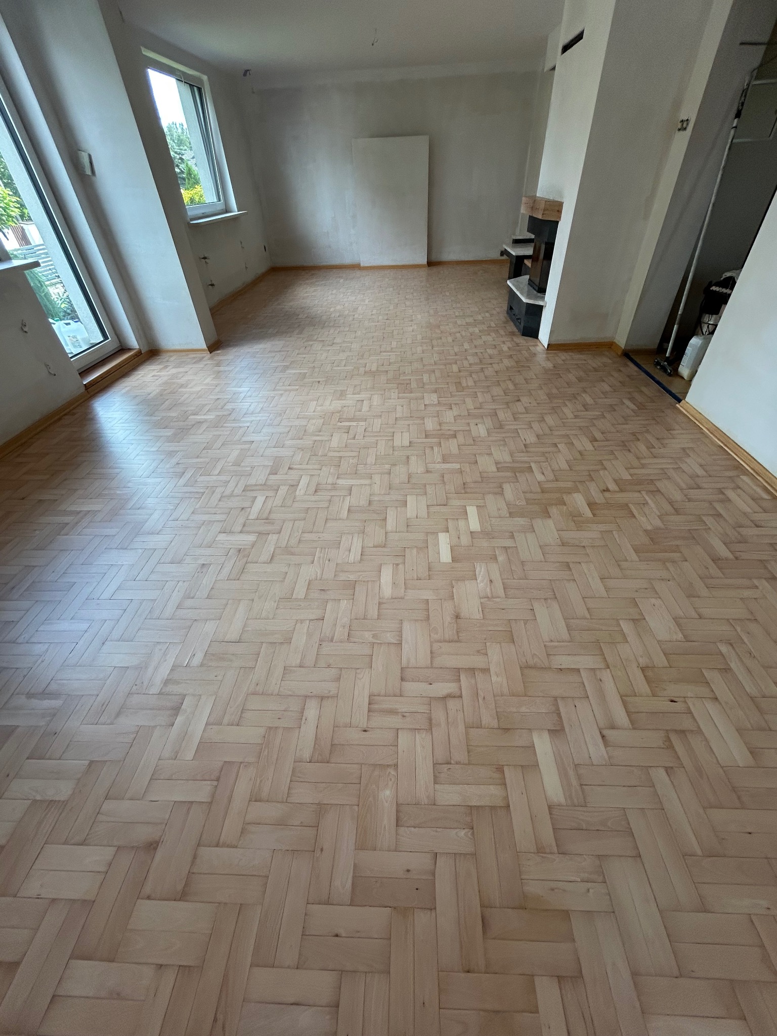 Jasna, odnowiona podłoga parkietowa ułożona we wzór jodełki w przestronnym, jasnym pokoju z oknami. Ściany przygotowane do malowania, widoczny kominek.