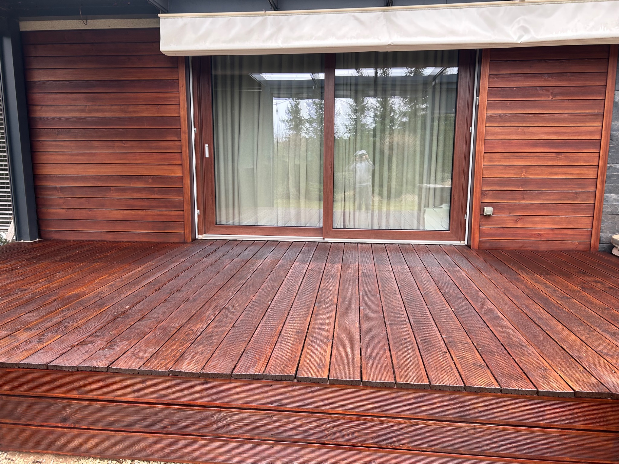 Impregnowany taras z drewna egzotycznego, deski ułożone poziomo, widoczne słoje drewna, brązowa kolorystyka, w tle przesuwne drzwi tarasowe i fragment elewacji.