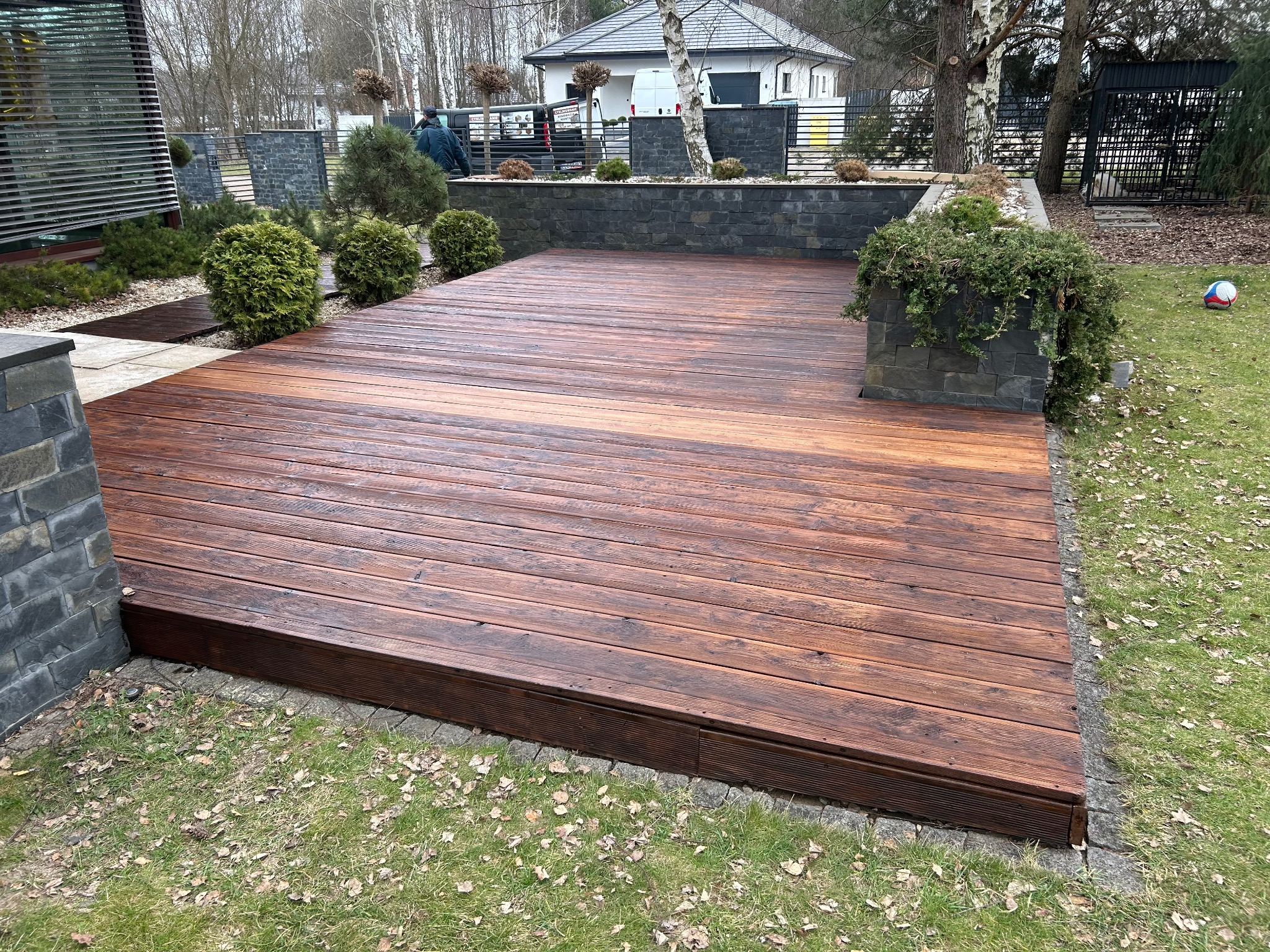 Impregnowany taras z desek na zewnątrz, otoczony zielenią i elementami kamiennymi. Widoczne detale wykończenia i impregnacji drewna. Ujęcie z niskiej perspektywy.