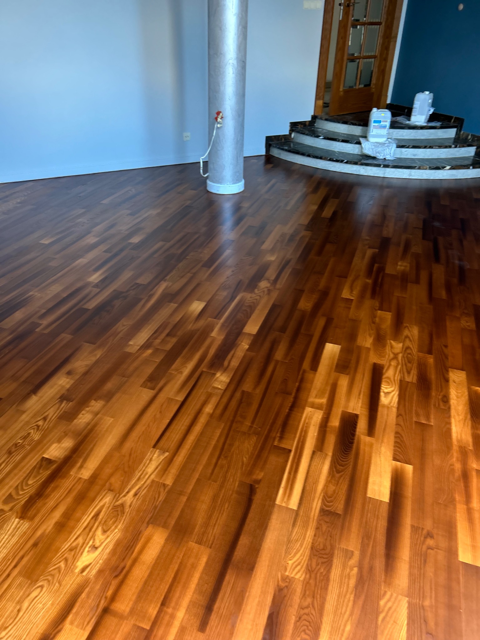 Błyszcząca, odnowiona drewniana podłoga parkietowa w jasnym odcieniu brązu, z widoczną kolumną i schodami w tle. Widoczne ślady po cyklinowaniu.