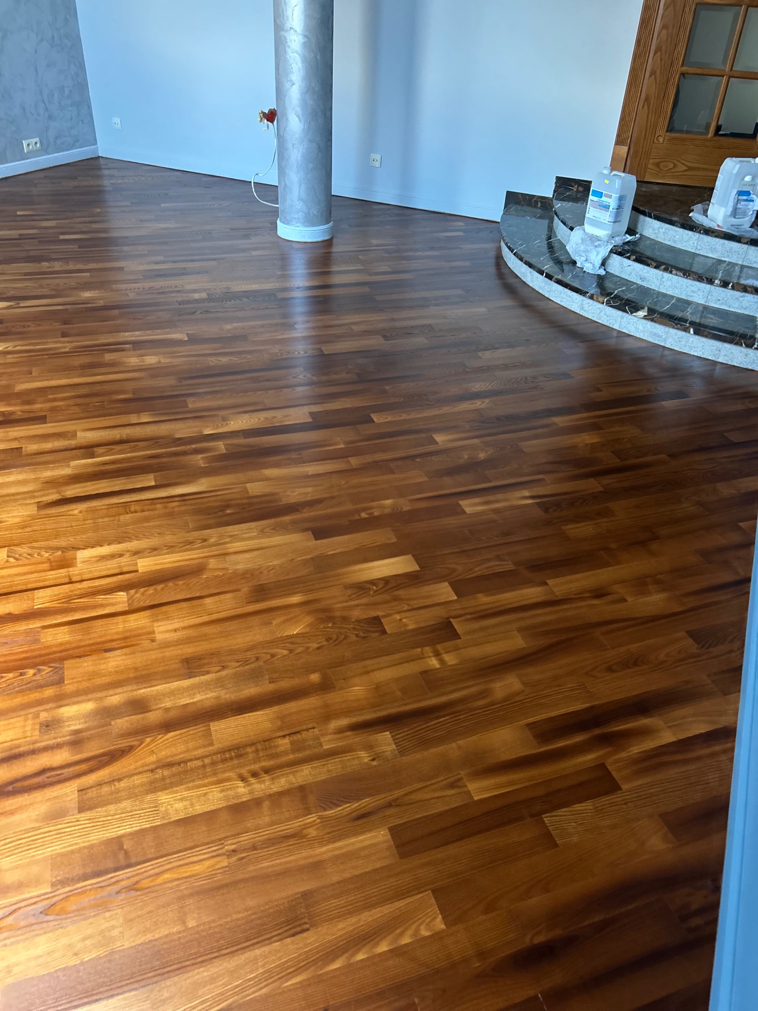 Błyszcząca, świeżo wycyklinowana podłoga parkietowa z wyraźnym usłojeniem, widoczna kolumna i schody z ciemnego marmuru. W tle białe ściany i drewniane drzwi.