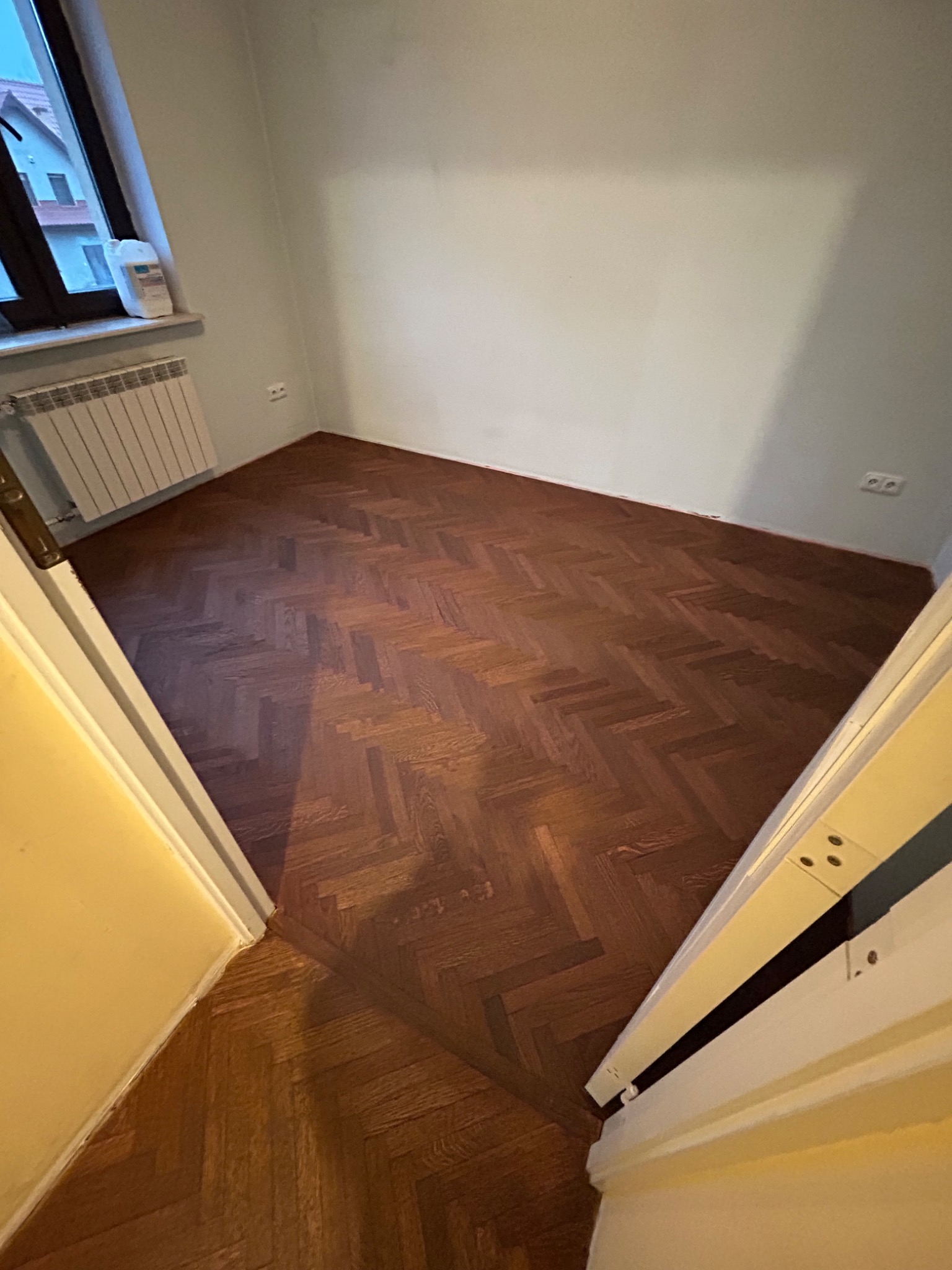Ciemnobrązowy parkiet ułożony w jodełkę w pokoju z grzejnikiem pod oknem. Widok zza progu, ściany pomalowane na jasny kolor.