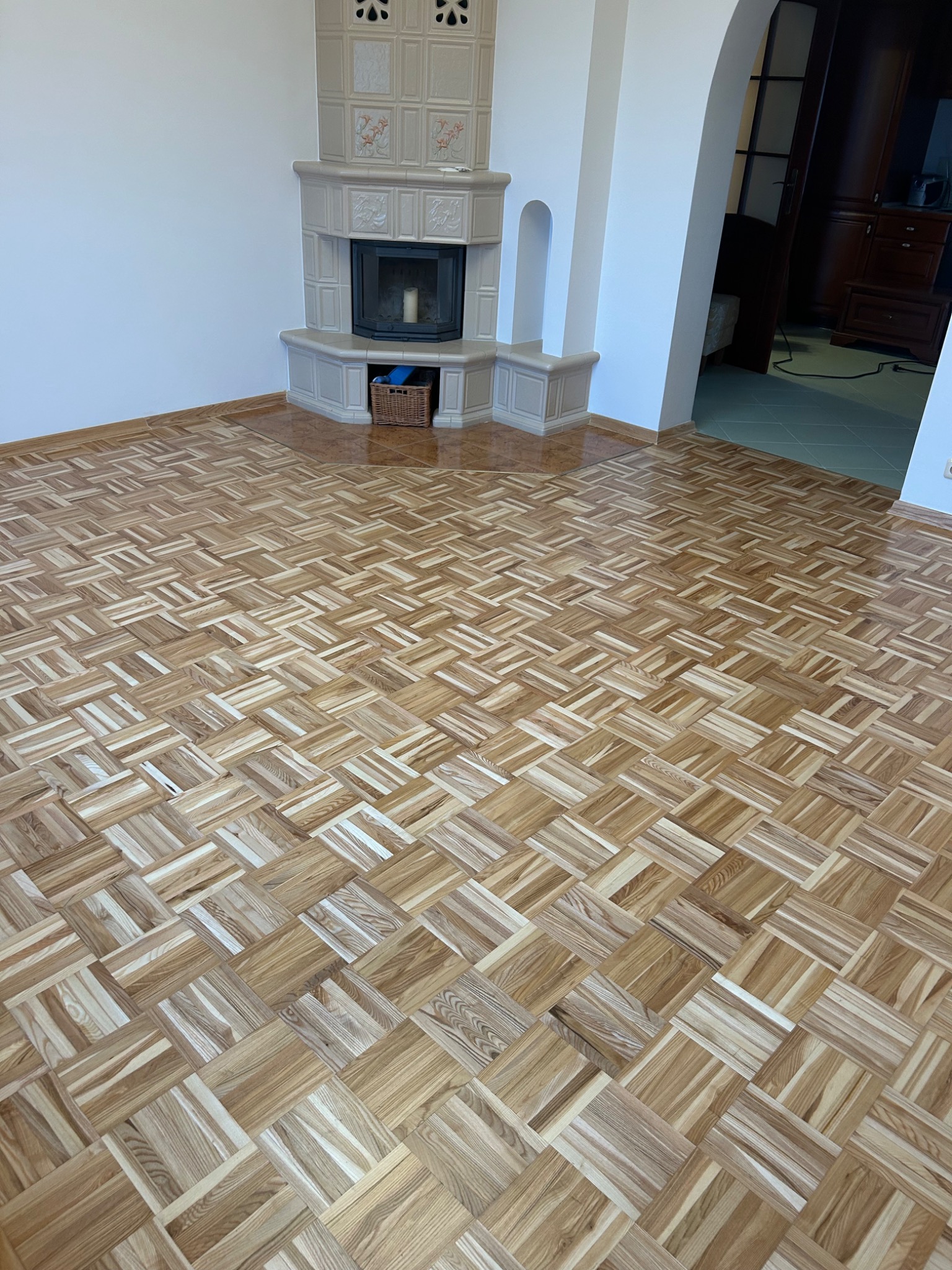 Parkiet ułożony w kwadraty, jasne drewno, widok na kominek z dekoracyjnymi kaflami i wejście do innego pomieszczenia. Jasne wnętrze po renowacji.