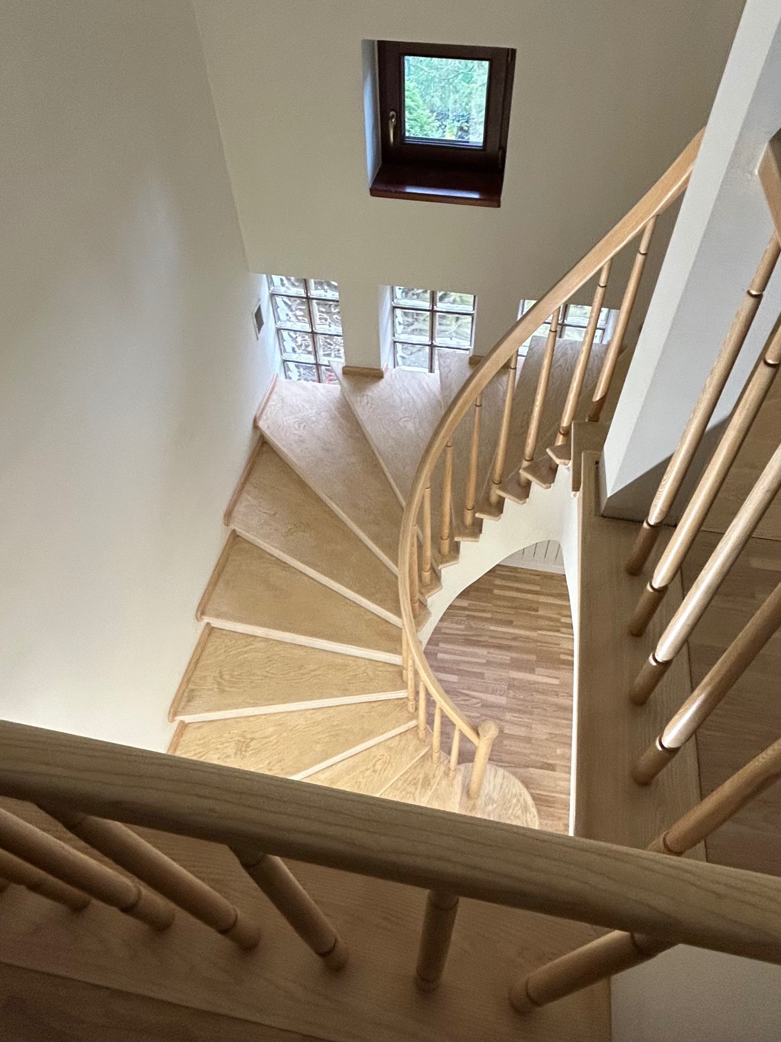 Drewniane, kręte schody z jasnego drewna, widziane z góry. Stopnie wykończone, balustrada z okrągłych elementów. W tle okna z luksferami i małe okno.