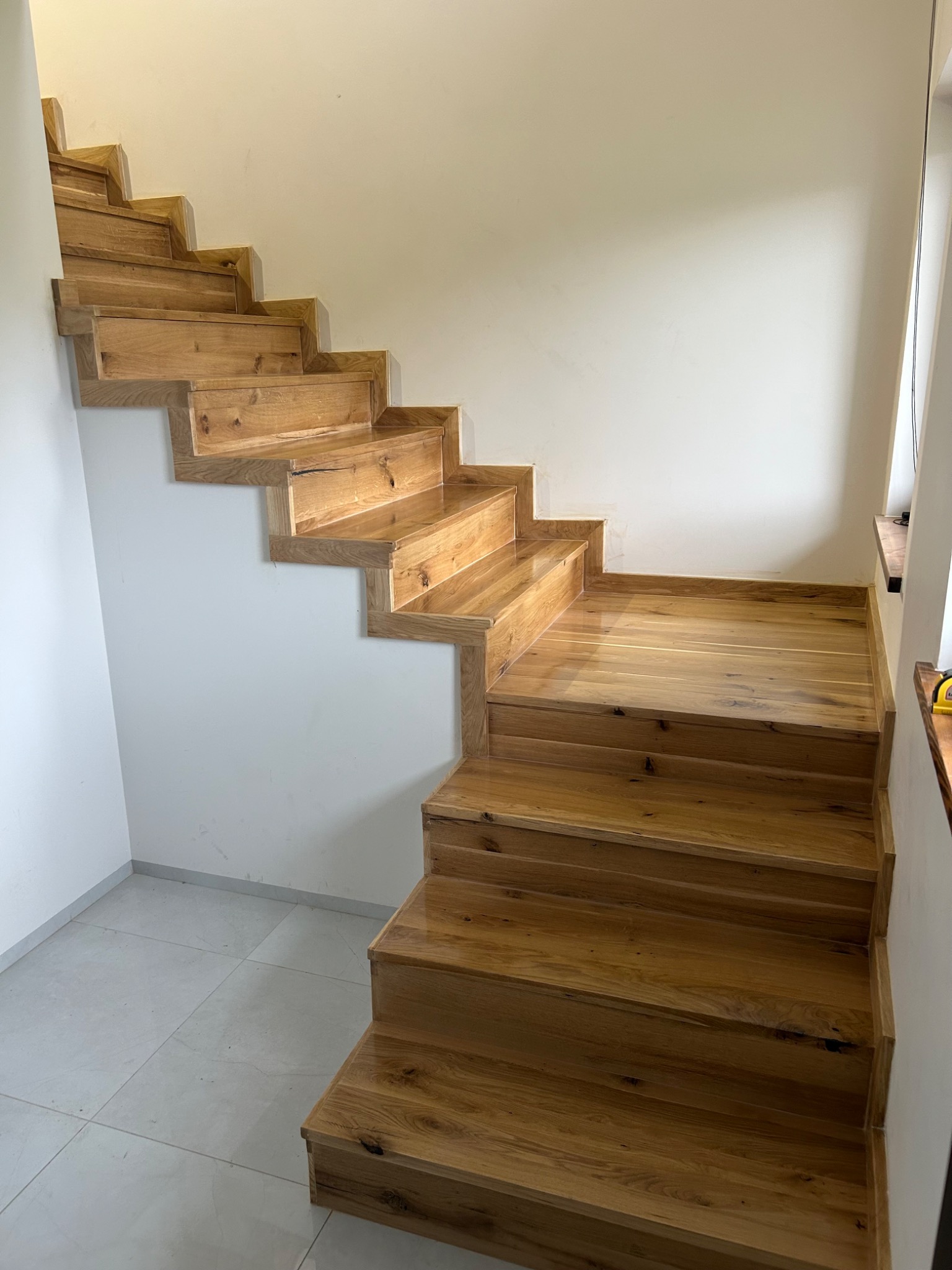 Drewniane schody dębowe, lakierowane, z widocznymi słojami drewna, zamontowane przy białej ścianie. Stopnie schodów o nieregularnym kształcie, widok z dołu.