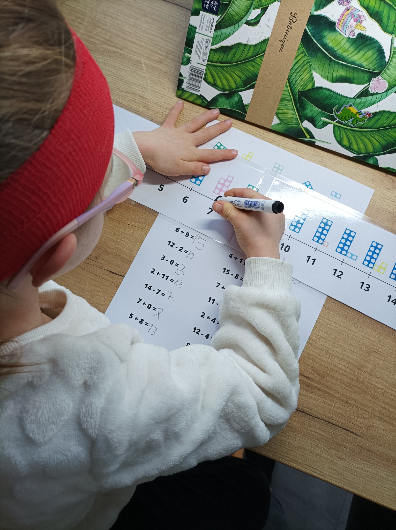 Dziecko w czerwonej opasce rozwiązuje zadania matematyczne na kartce z pomocą linijki z zaznaczonymi liczbami i polami do zaznaczania, obok leży pudełko z motywem liści.