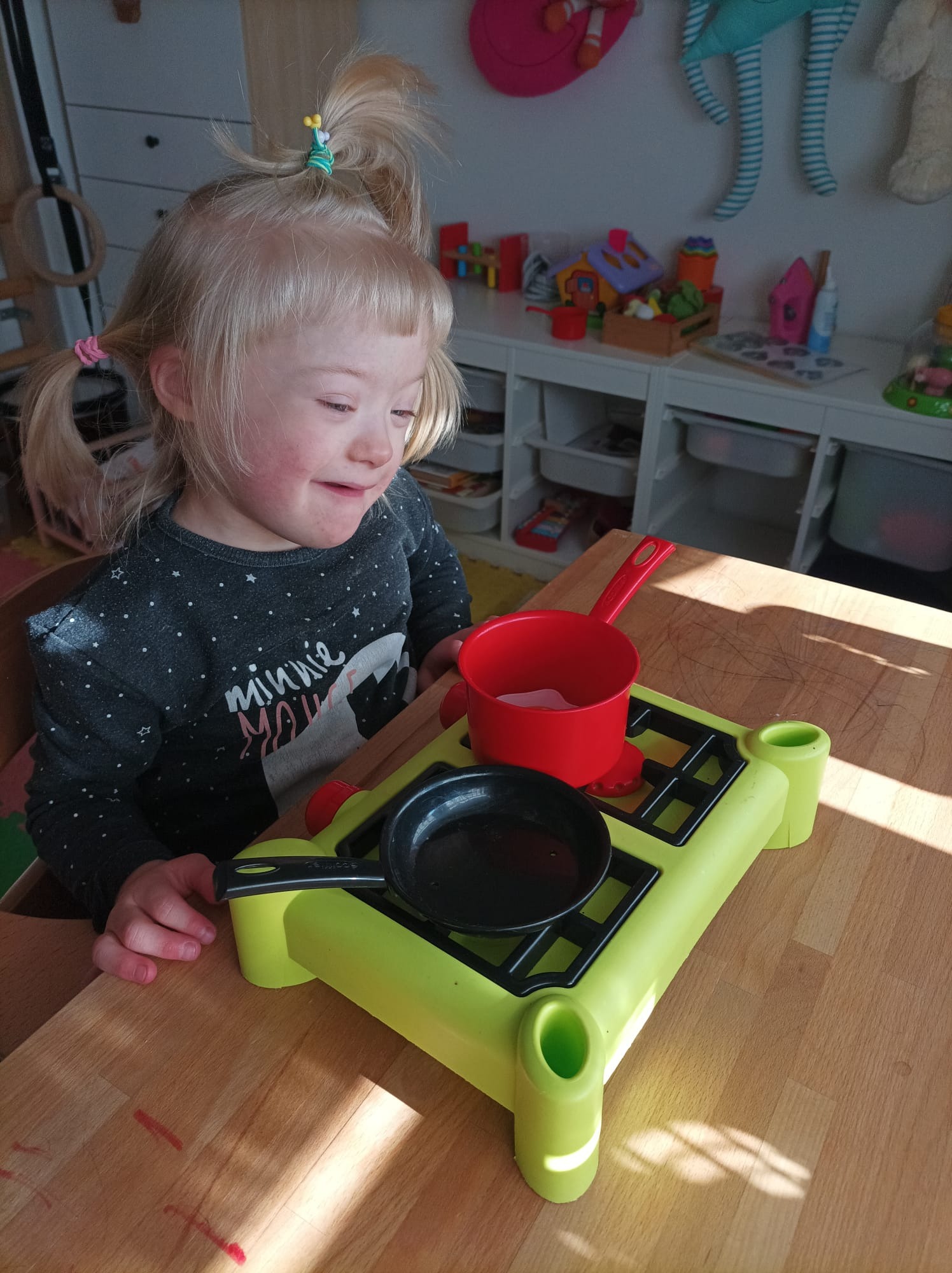 Dziewczynka z zespołem Downa bawi się zabawkami kuchennymi: zieloną kuchenką, czarną patelnią i czerwonym garnkiem na drewnianym stole, w tle zabawki na półkach.