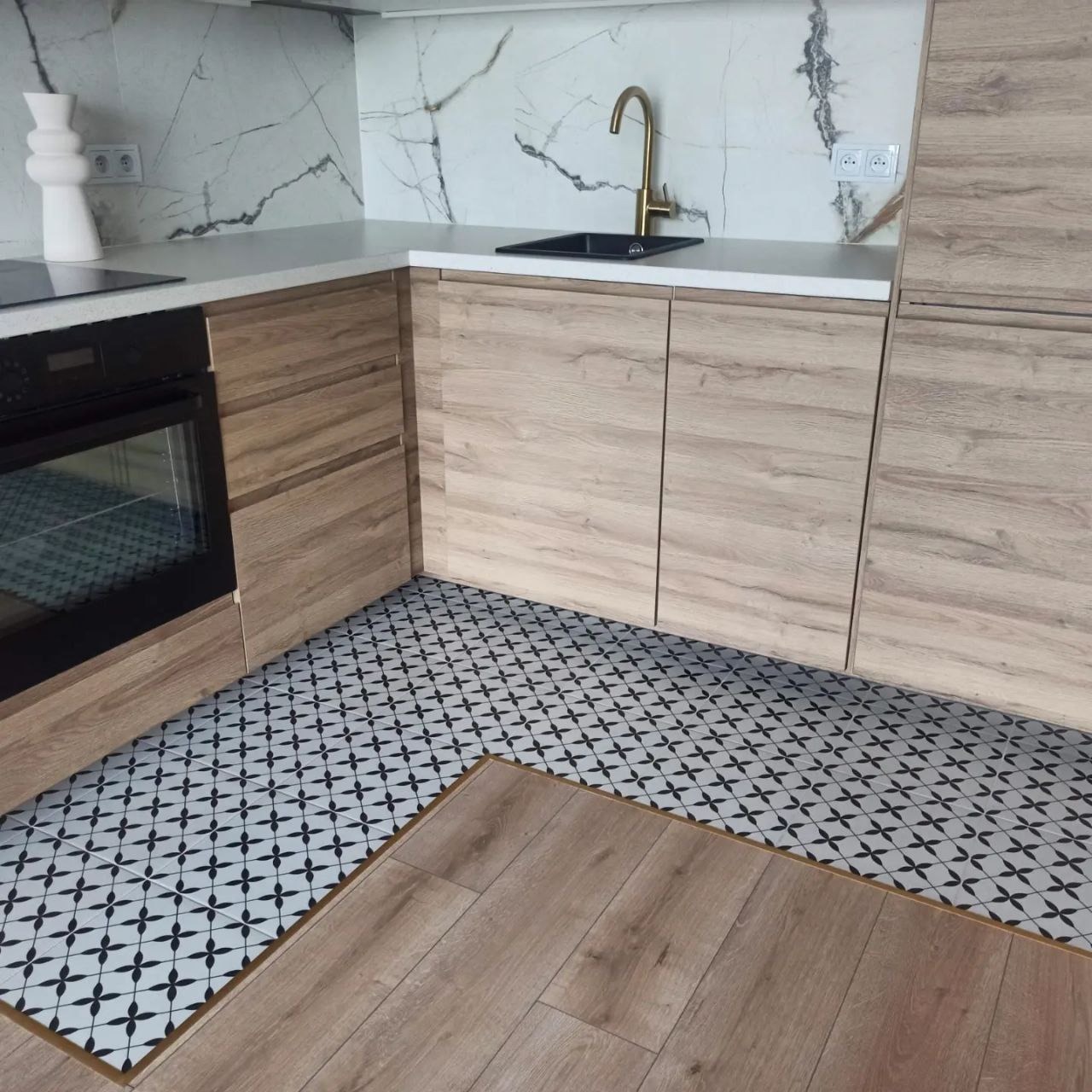Nowoczesna kuchnia w Warszawie z drewnianymi szafkami, marmurowym blatem, złotą armaturą i wzorzystą podłogą, łącząca panele z płytkami.