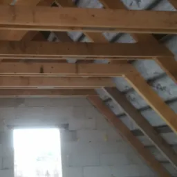 Konstrukcja drewniana dachu poddasza w trakcie budowy, widoczne krokwie i mur z pustaków, w tle szara izolacja i okno z widokiem na zewnątrz w jasnym świetle.