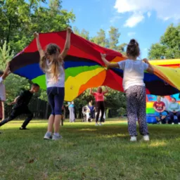 Grupa dzieci trzymająca nad głowami wielobarwną płachtę w kształcie spadochronu, w tle dmuchany zamek i siedzący dorośli.