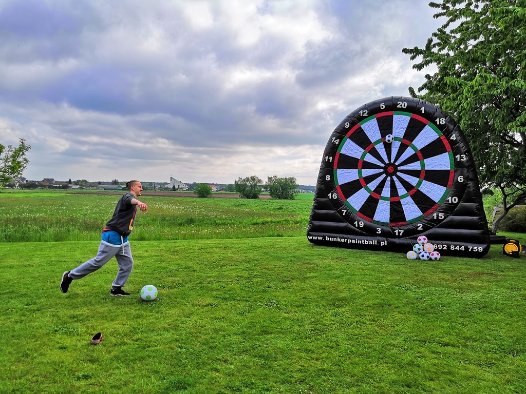 Chłopiec kopie piłkę w kierunku dmuchanej tarczy do darta na trawiastym terenie; w tle widoczne zabudowania wiejskie pod pochmurnym niebem.