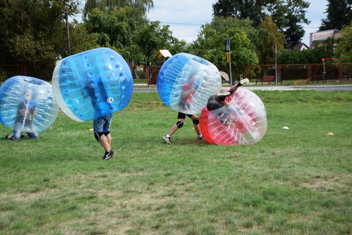 Grupa osób w przezroczystych kulach na trawiastym terenie, w tle drzewa i budynki, zabawa w kulach na świeżym powietrzu.