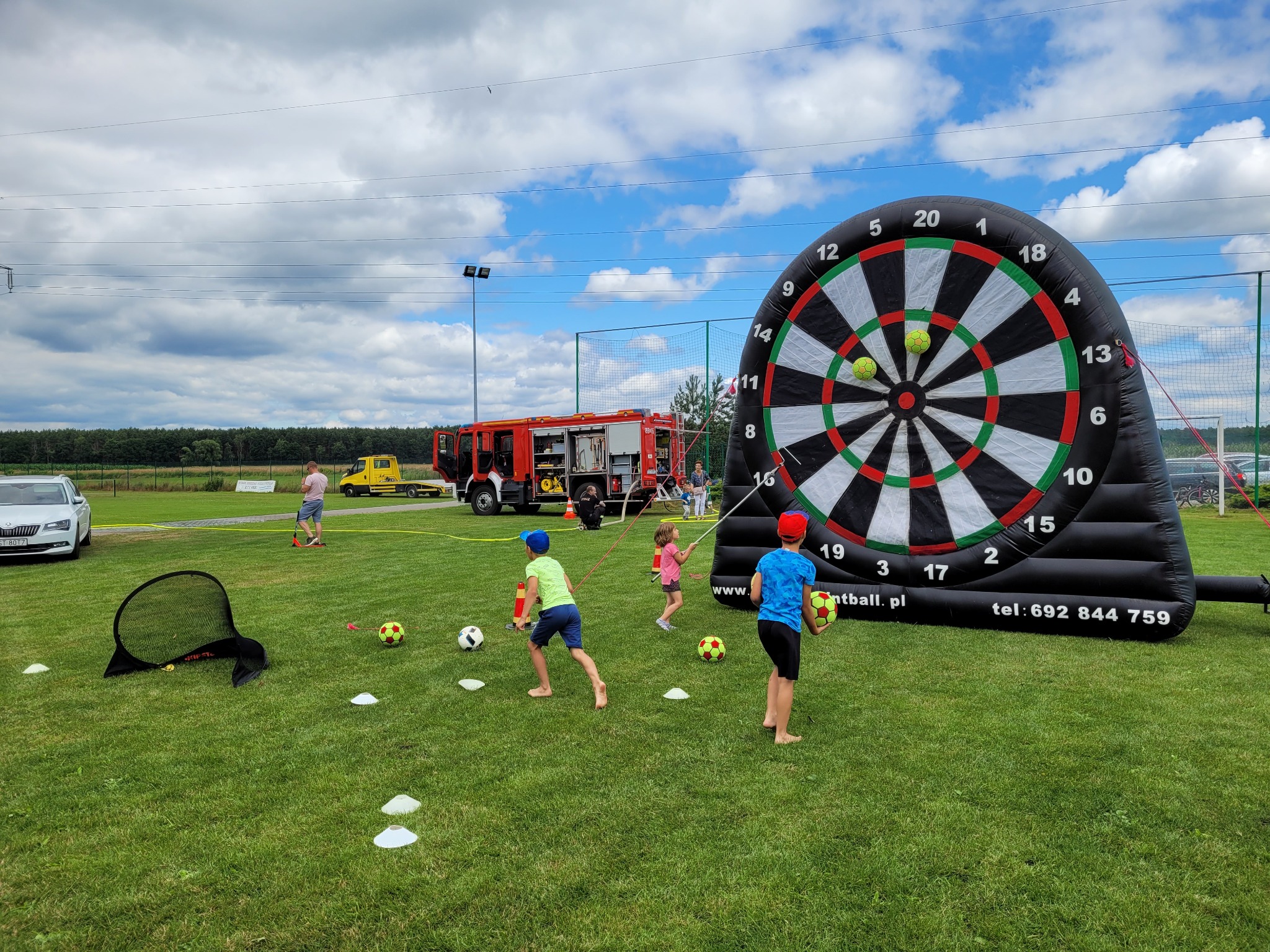 Dzieci grają w dmuchane piłkarskie rzutki na trawie, w tle wóz strażacki i inne osoby, na zielonym trawniku leżą piłki i mała bramka.