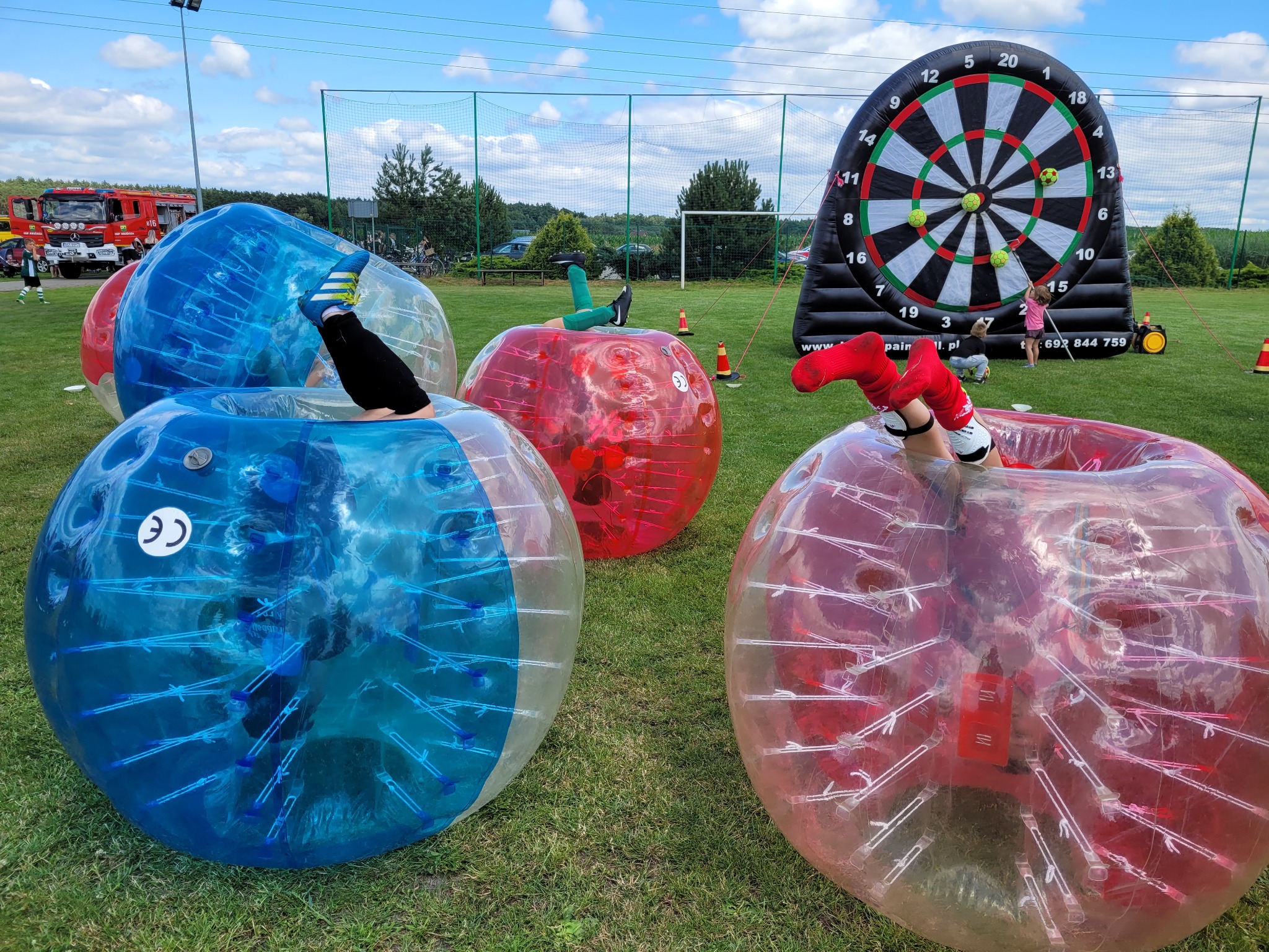Kolorowe, przezroczyste kule do zabawy na trawie, w tle dmuchana tarcza do rzutek i wóz strażacki.