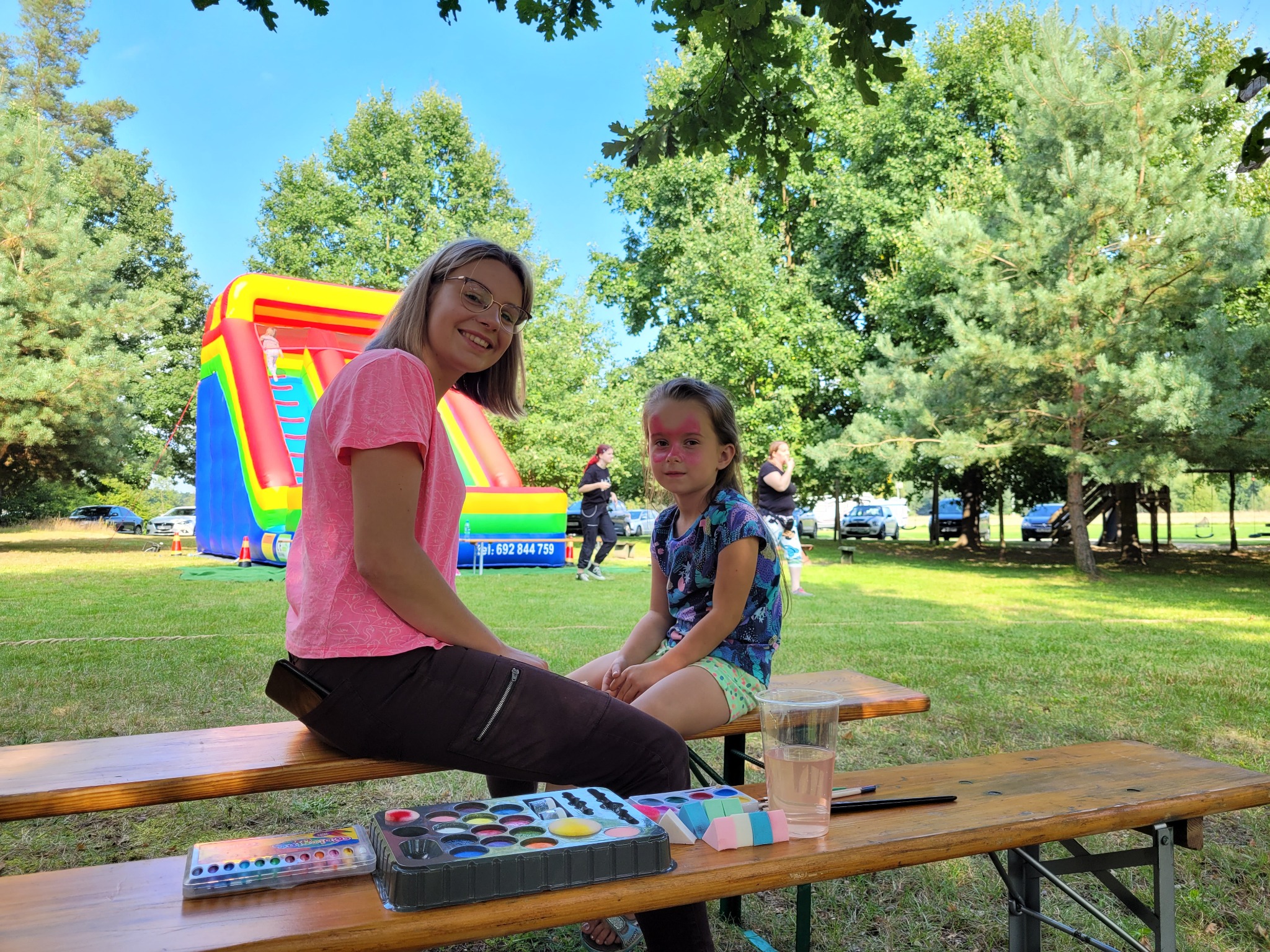 Kobieta z paletą farb do twarzy siedzi na ławce obok dziewczynki z pomalowaną twarzą; w tle dmuchana zjeżdżalnia i osoby na trawie.
