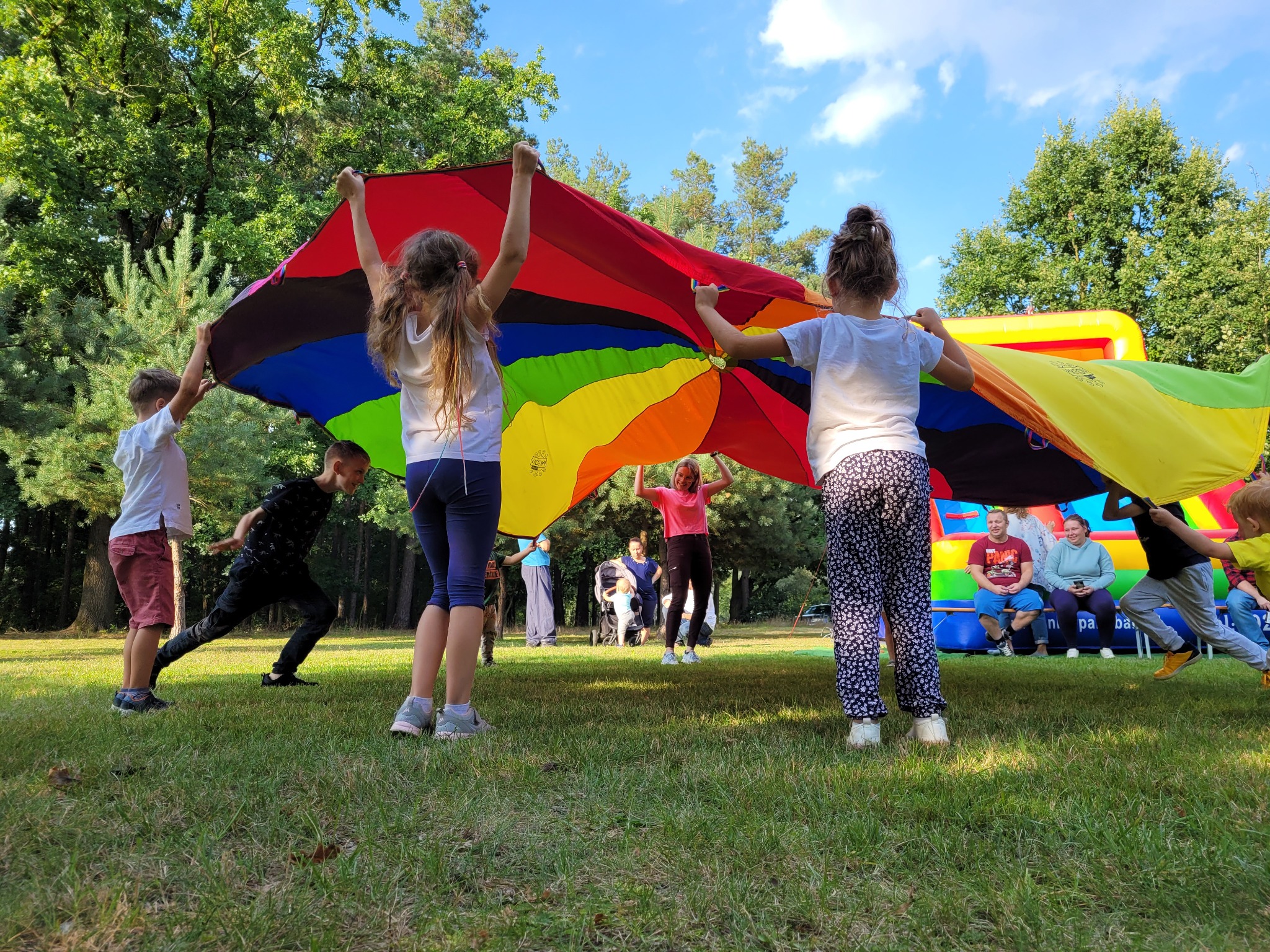 Grupa dzieci trzymająca nad głowami wielobarwną płachtę w kształcie spadochronu, w tle dmuchany zamek i siedzący dorośli.
