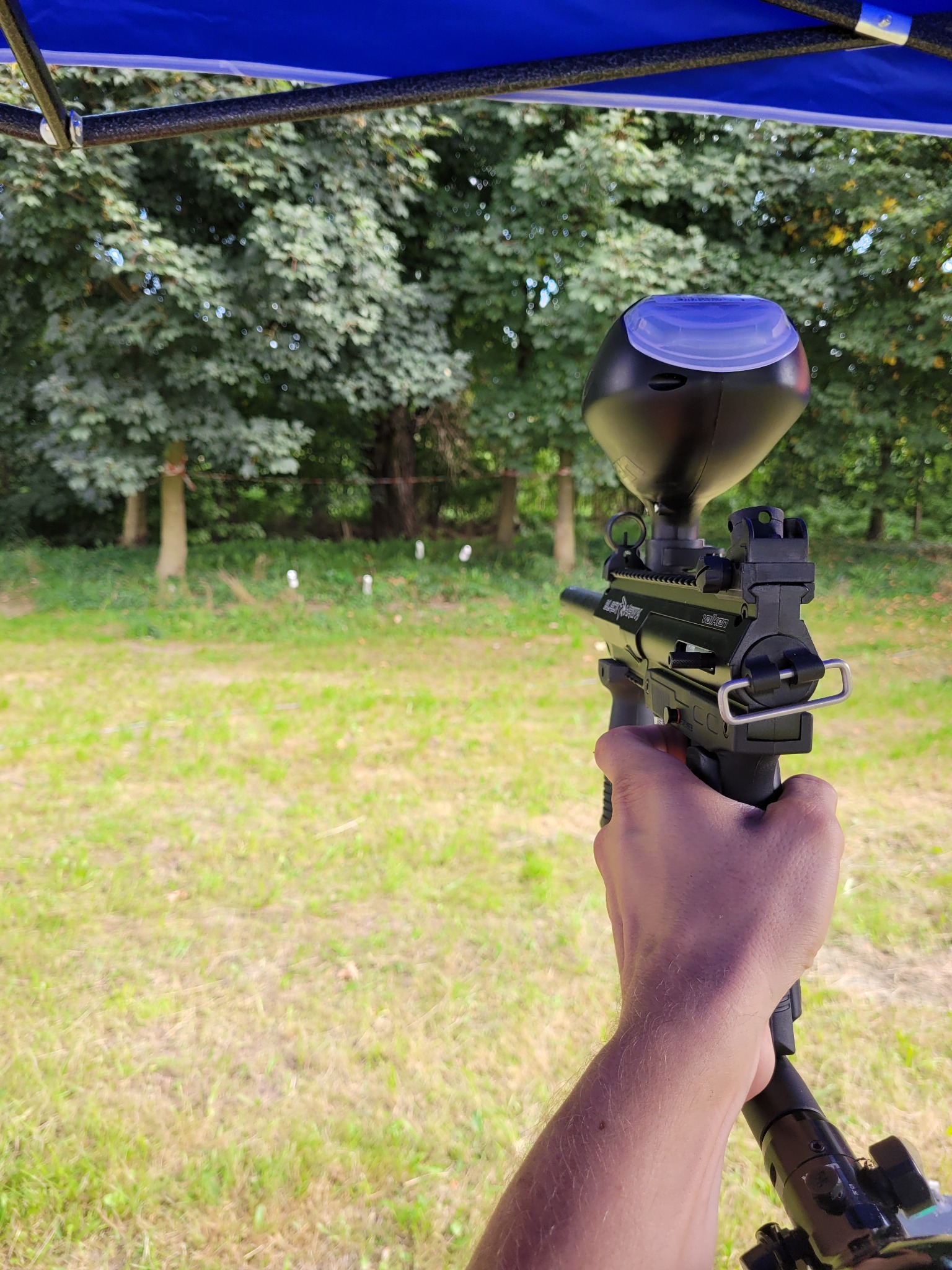 Osoba trzyma w dłoni karabinek do paintballa na tle zielonego trawnika i drzew, widoczne białe ślady po kulkach.