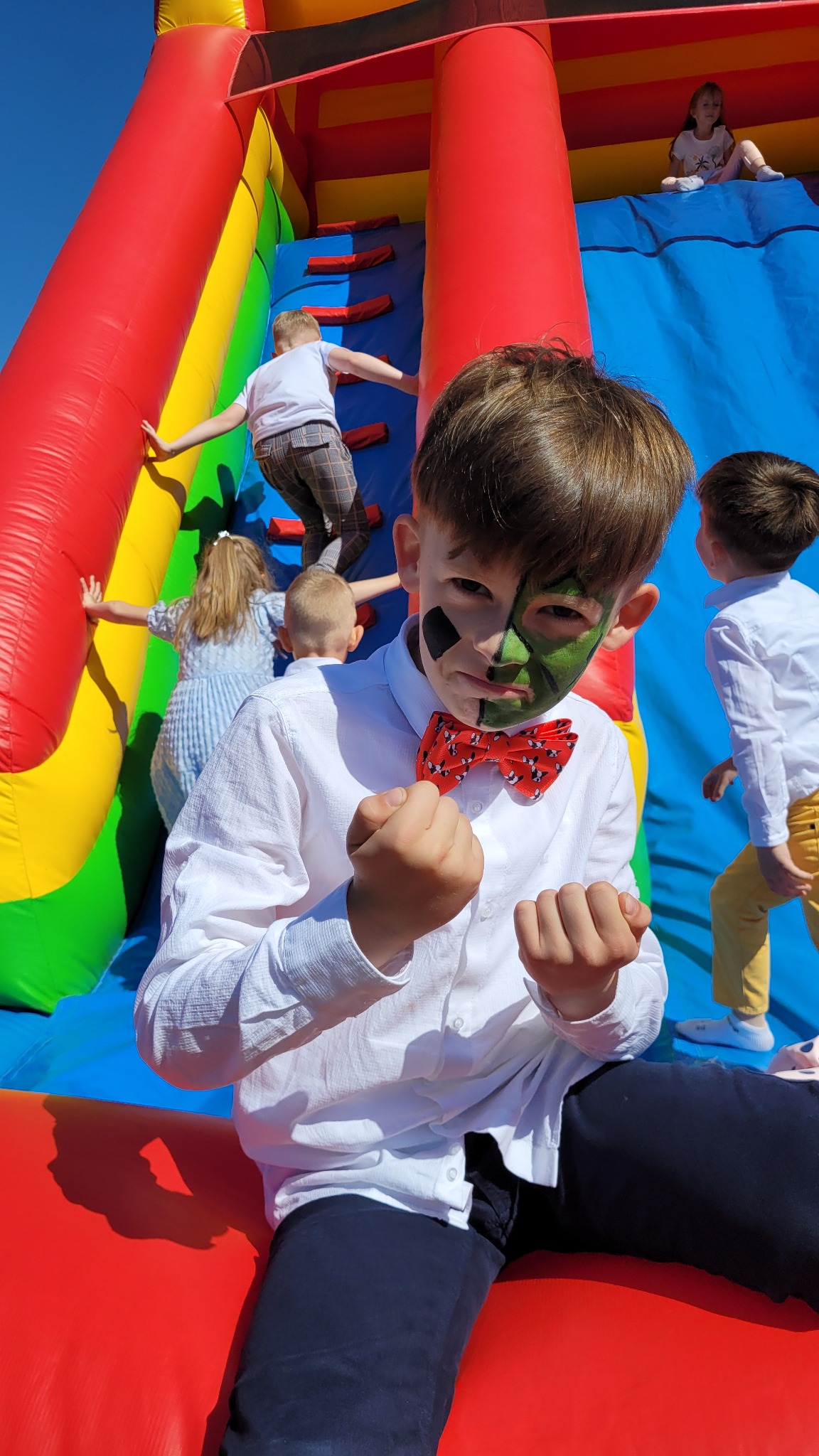 Chłopiec z pomalowaną twarzą w stylu superbohatera i muchą w grochy, na tle kolorowej, dmuchanej zjeżdżalni z innymi dziećmi.