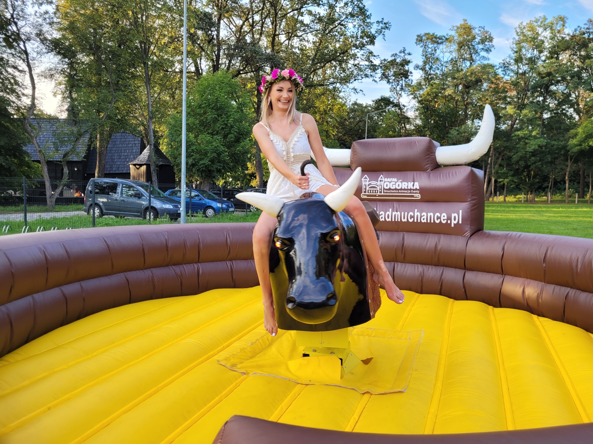 Uśmiechnięta blondynka w wianku na głowie siedzi na mechanicznym byku, który znajduje się na dmuchanej arenie w plenerze.