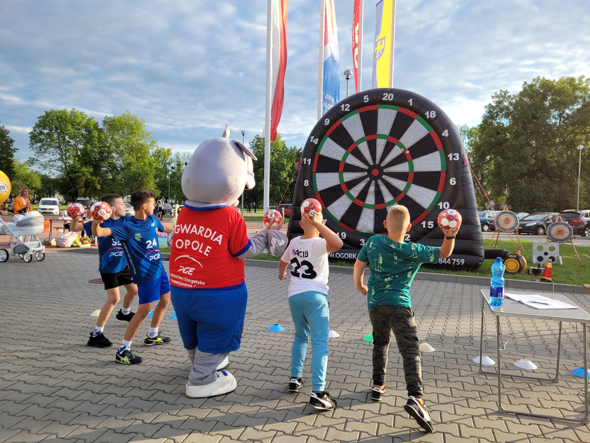 Uczestnicy zabawy rzucają piłkami do tarczy dmuchanej podczas eventu plenerowego z animatorem w stroju słonia i flagami w tle.